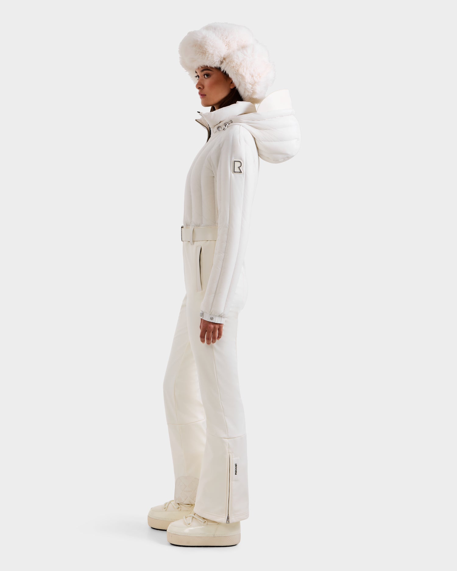 A woman in the Rudsak SAGE - 8124782 Iceland fitted white ski suit, belted at the waist, stands in profile with a fluffy white hat and platform boots against a plain light background.