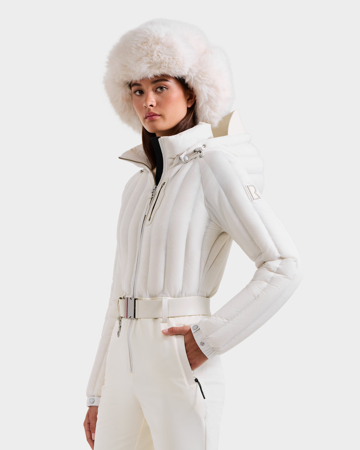 A woman poses against a plain light background, wearing the Rudsak SAGE - 8124782 Iceland stylish white belted women's ski suit paired with a large fluffy white fur hat.