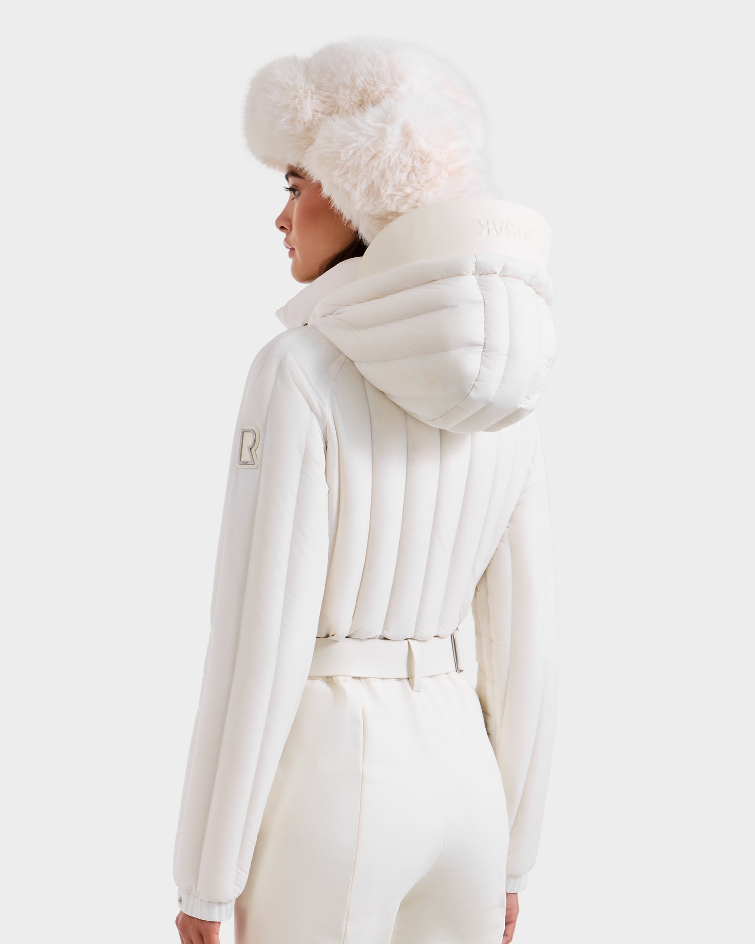 A woman models the Rudsak SAGE - 8124782 Iceland ski suit, featuring a belted jacket with quilted panels and hood, plus a fur hat. The simple background draws attention to this chic all-white winter look from Rudsak.