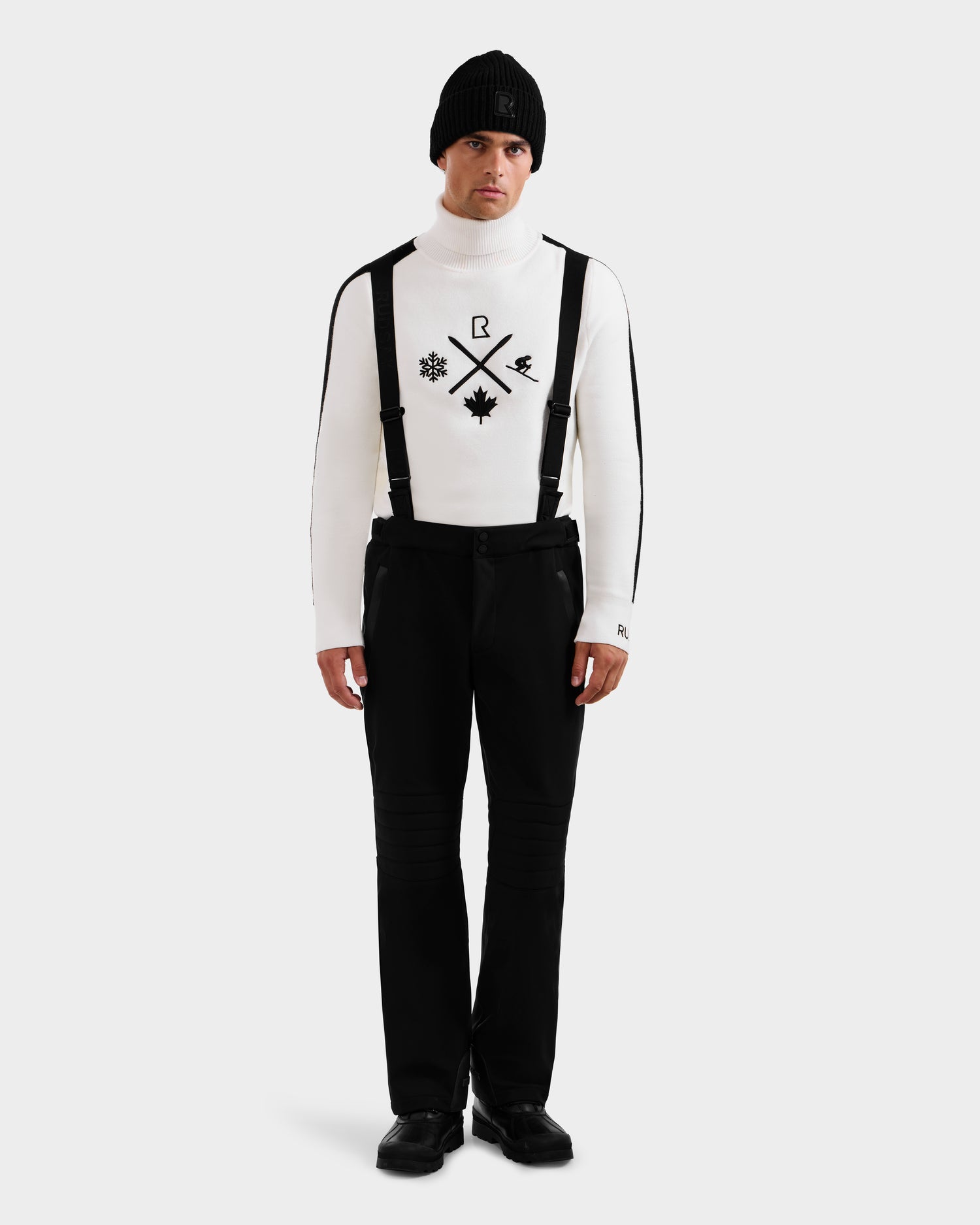 A man stands facing forward against a plain white background, wearing the Rudsak SAMPSON - 6125586 Black beanie, paired with a white turtleneck featuring black ski-themed symbols, black suspenders, black pants, and black shoes.