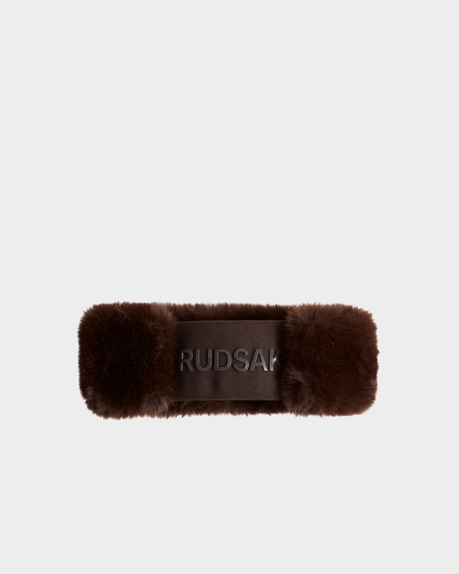 The SANDIE - L425552 Espresso by Rudsak is a brown faux fur headband featuring "RUDSAK" embroidered on a sleek front panel, ideal for enhancing any winter look. Shown against a simple light background.