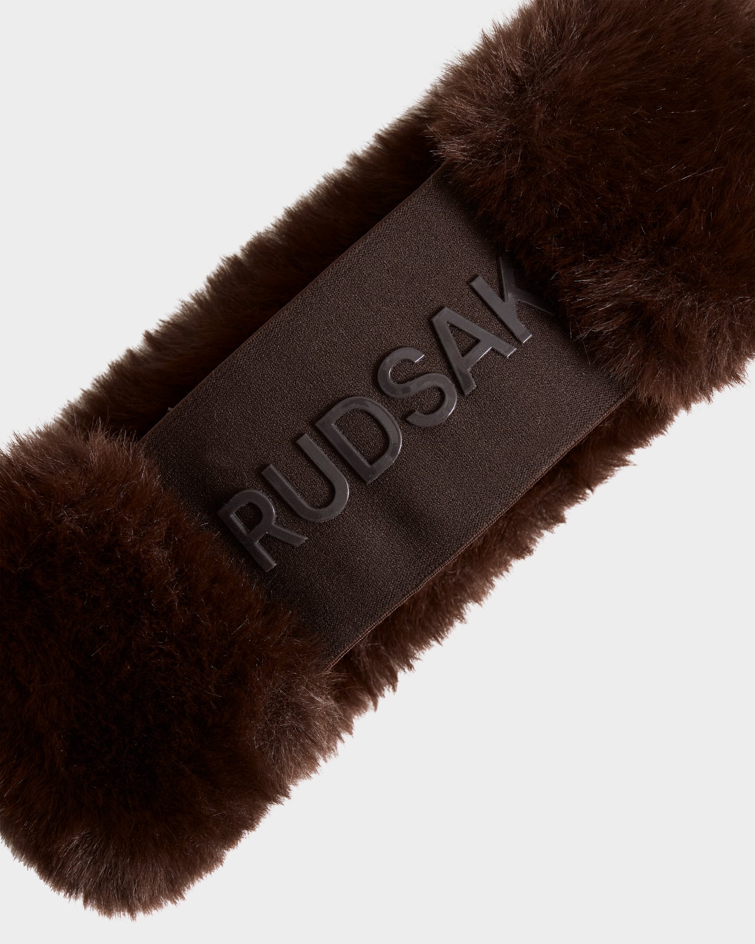 A close-up of the SANDIE - L425552 Espresso faux fur headband by Rudsak, featuring the brand embossed on a matching fabric band—an essential winter accessory.