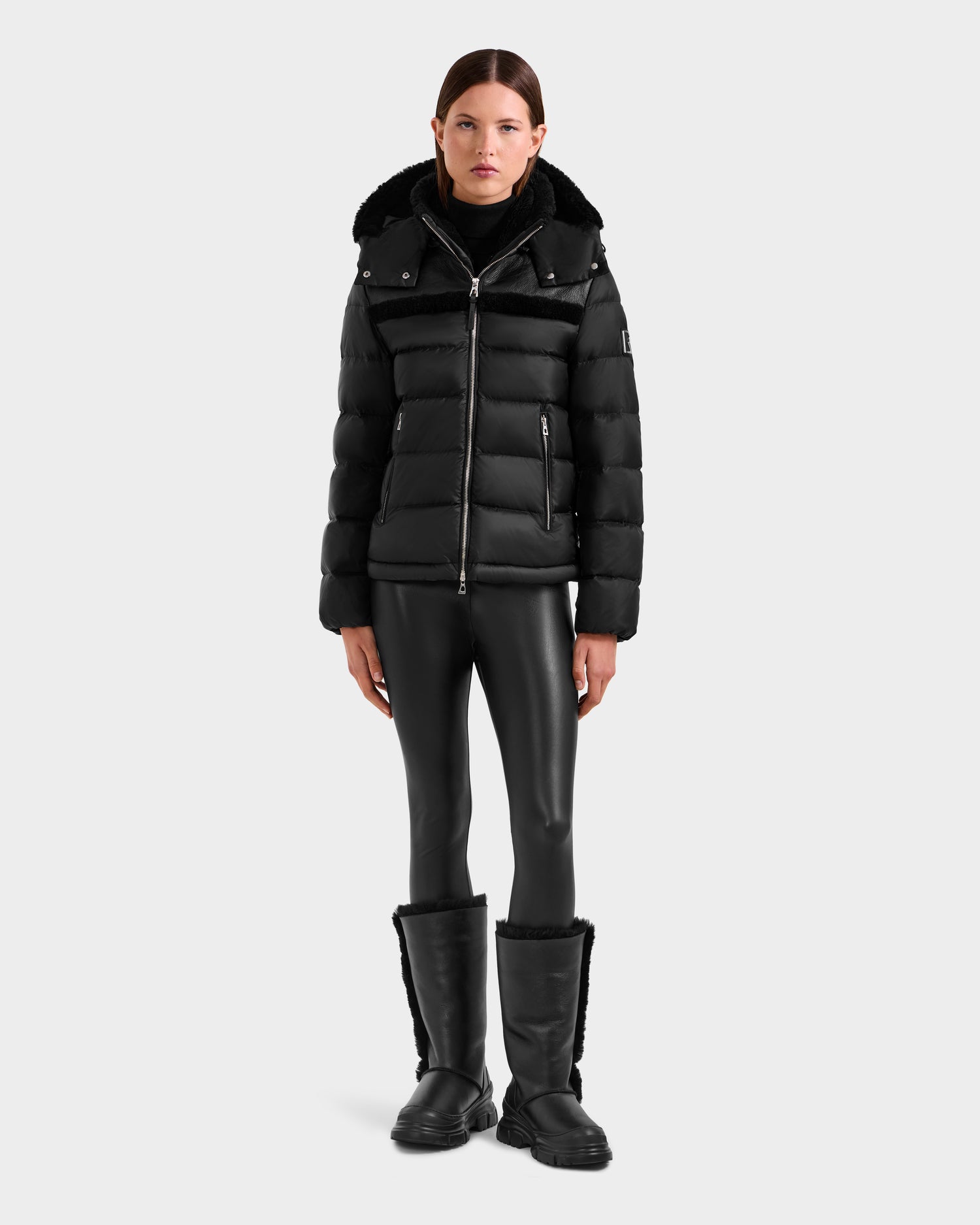 A person faces forward wearing the Rudsak SANDRA - 8125612 Black women’s shearling puffer jacket, paired with a black turtleneck, black leather-look leggings, and black knee-high fur-trim boots, set against a plain light gray background.