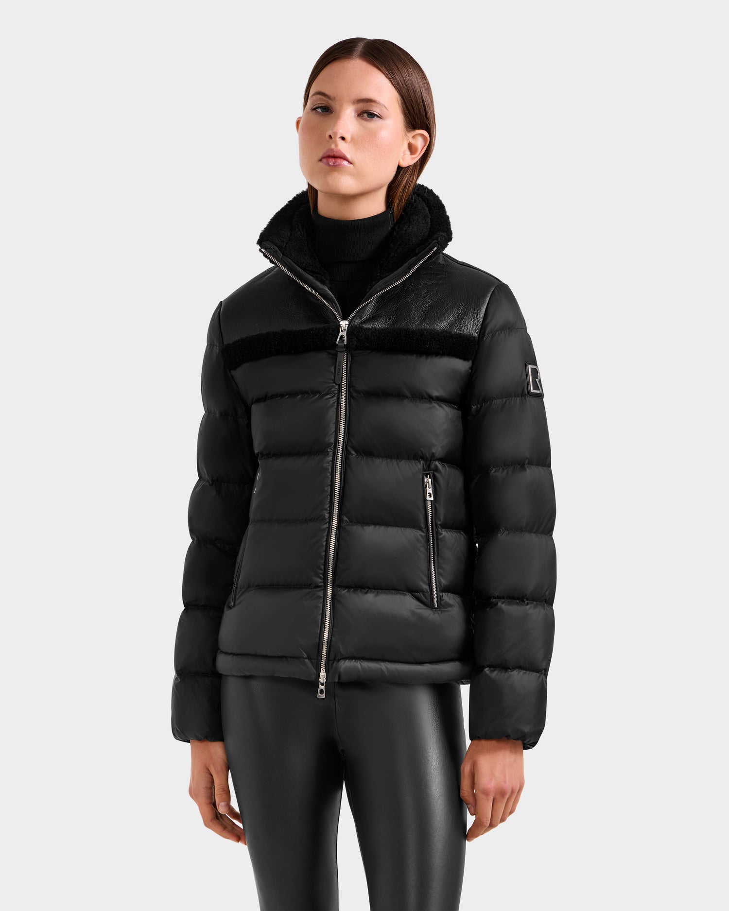A woman models the Rudsak SANDRA - 8125612 Black women’s shearling puffer jacket with lightweight insulation and a shiny black finish, styled with black leggings against a plain light grey background.
