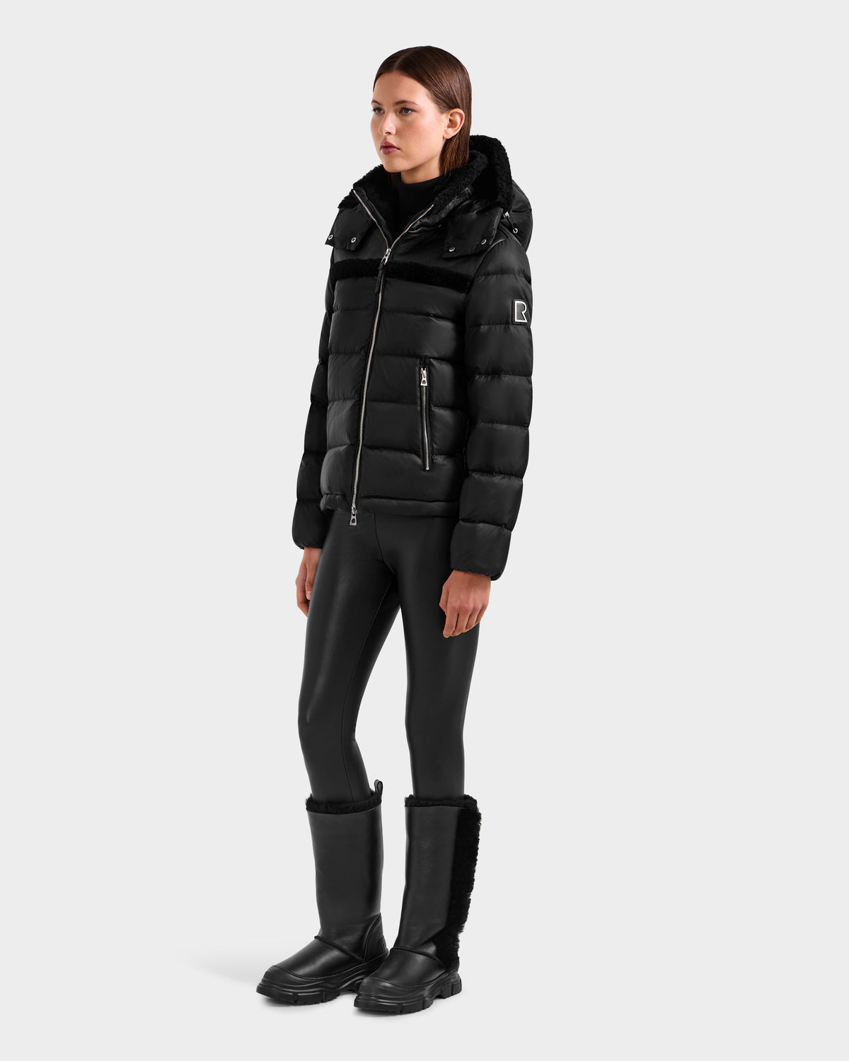 A woman with straight brown hair stands against a plain background in the Rudsak SANDRA - 8125612 Black women’s shearling puffer jacket, paired with black faux leather leggings and fur-trimmed mid-calf boots. Her hands are by her sides.