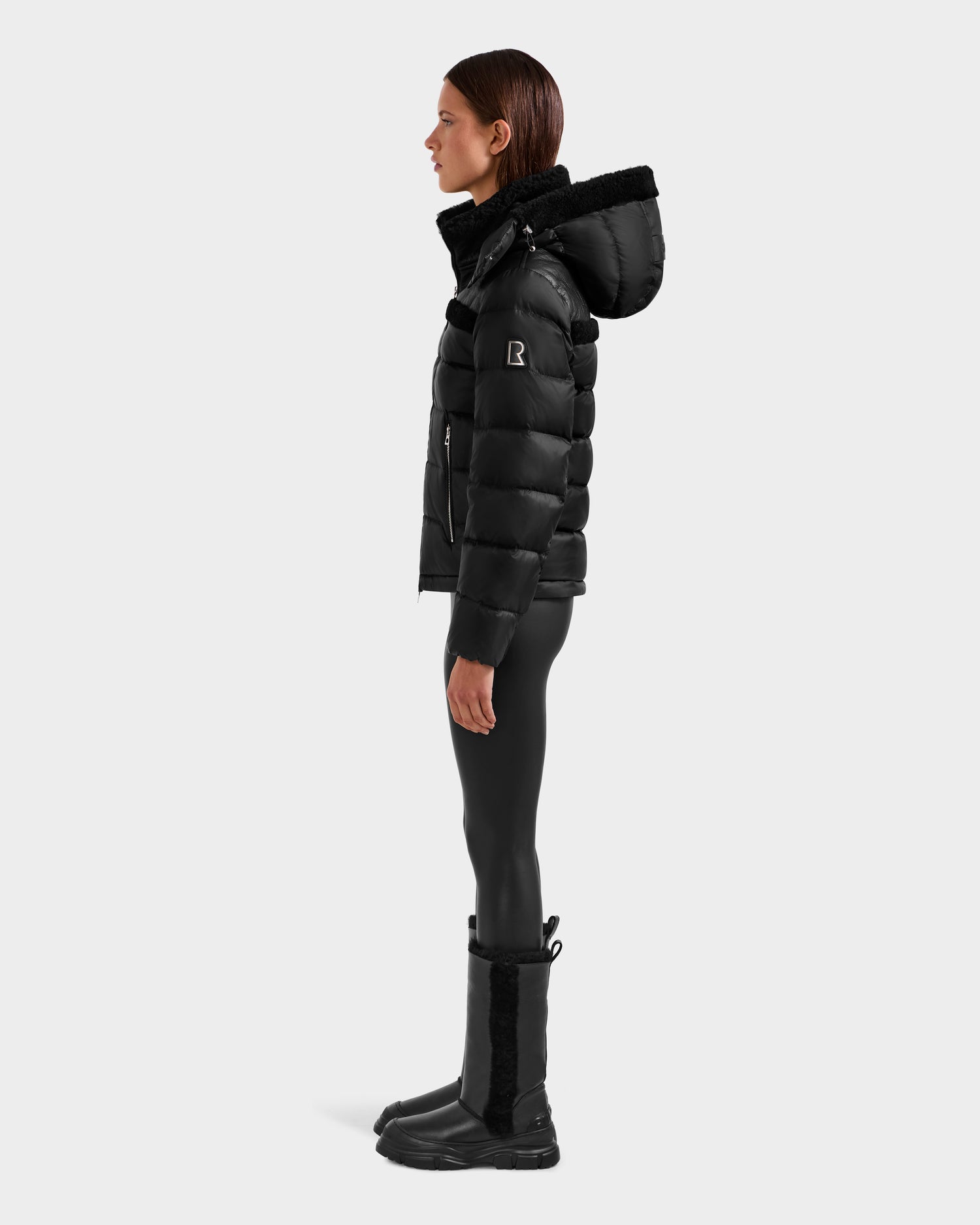 A person stands in profile wearing the Rudsak SANDRA - 8125612 Black women’s shearling puffer jacket with a hood, black leggings, and black boots against a plain light gray background for a modern look.