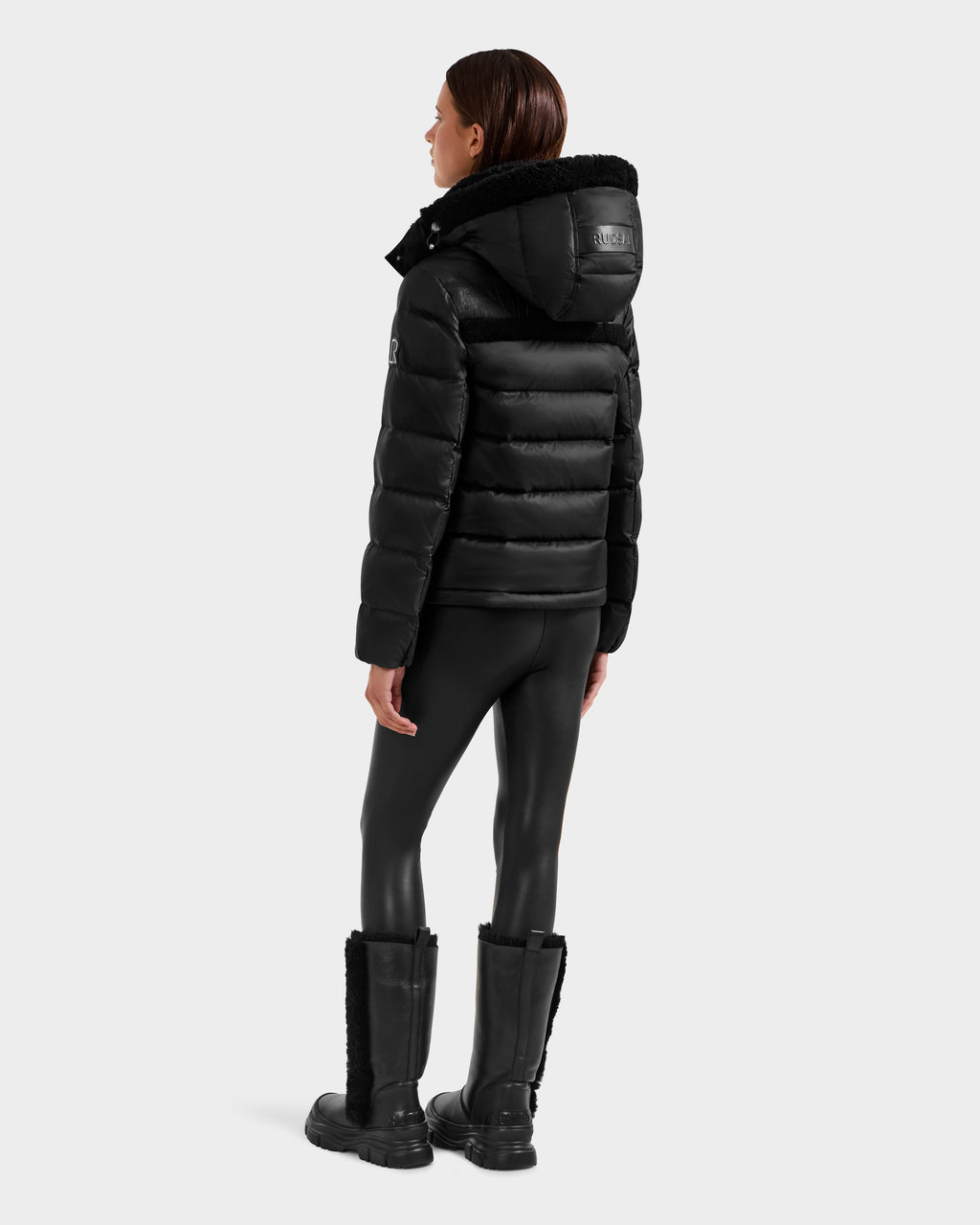 A person faces away, highlighting a modern look in Rudsak’s SANDRA - 8125612 Black women’s shearling puffer jacket with hood, paired with shiny black leggings and tall fur-trimmed black boots against a plain light backdrop.