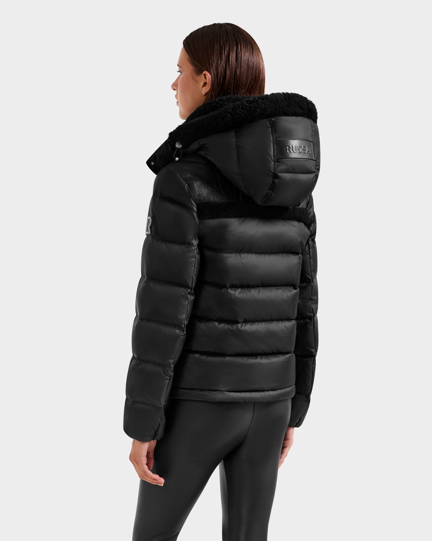 A person with straight brown hair wears the Rudsak SANDRA - 8125612 Black women's shearling puffer jacket and black leggings, shown from the back against a plain light gray background for a modern, stylish look.