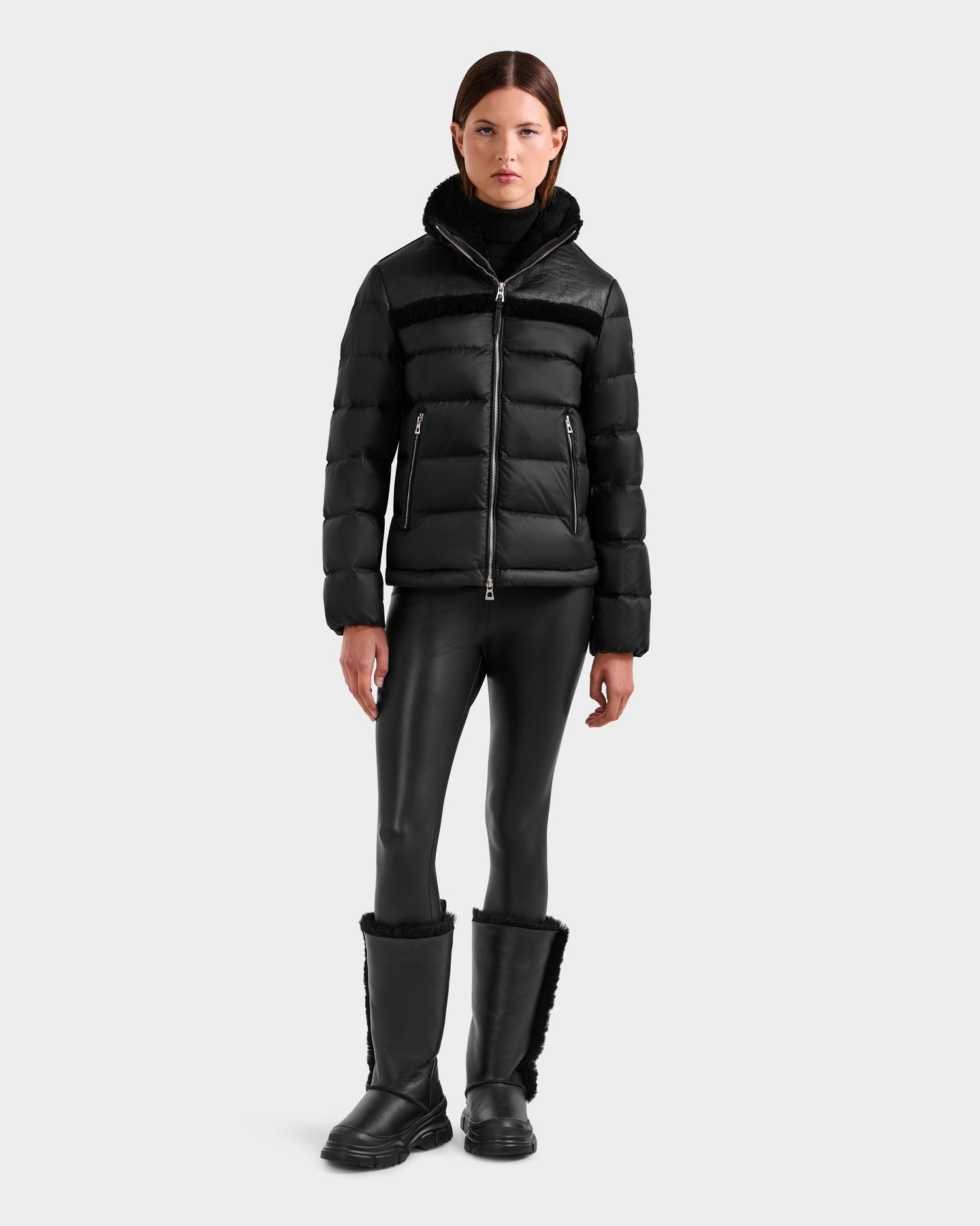 A woman gazes at the camera with a neutral expression, wearing the Rudsak SANDRA - 8125612 Black shearling puffer jacket, paired with black faux leather leggings and fur-trimmed boots for a modern look against a plain background.