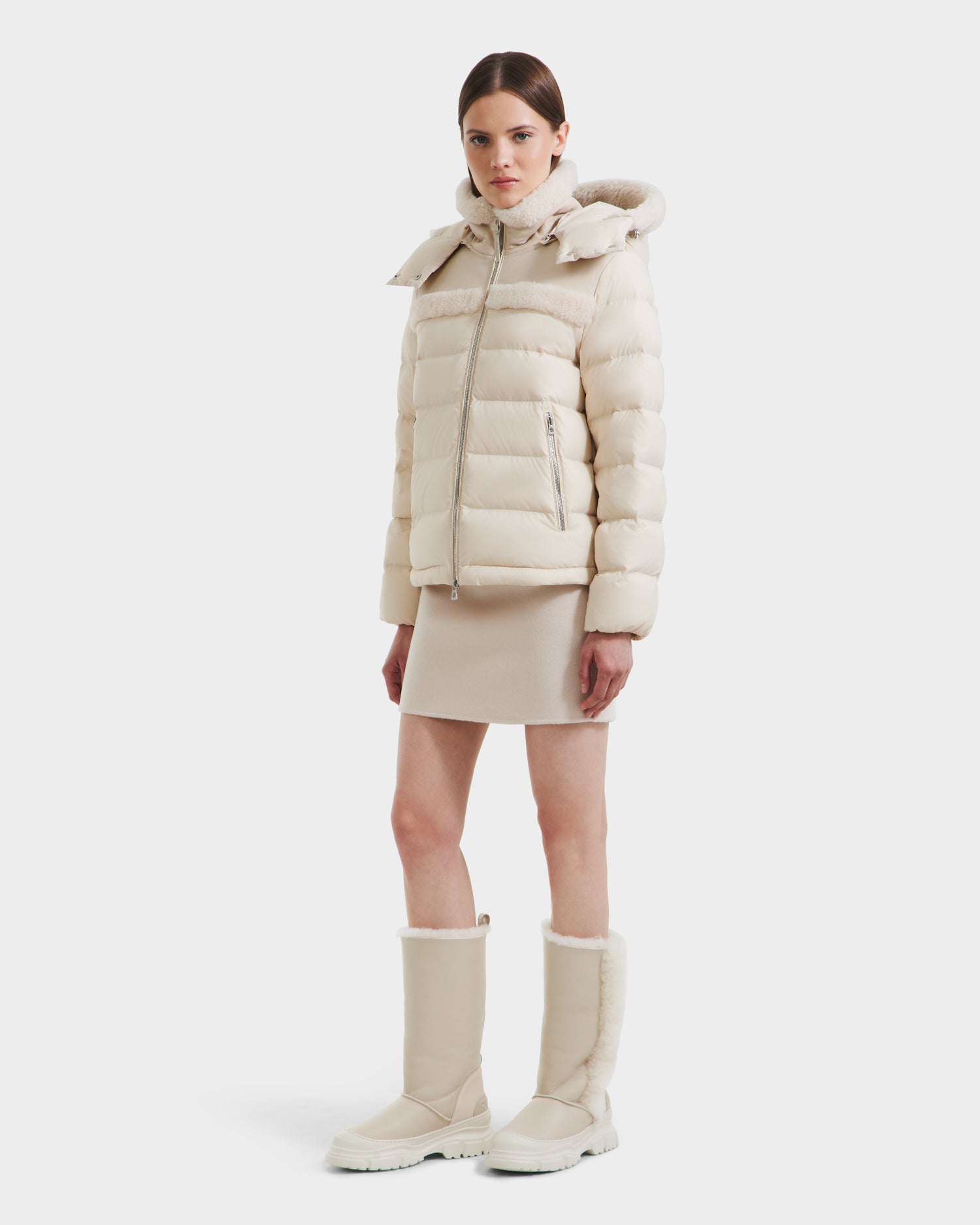 A woman stands facing forward in the Rudsak SANDRA - 8125612 Vanilla insulated winter coat with high collar and hood, a matching skirt, and knee-high beige boots against a plain white background.