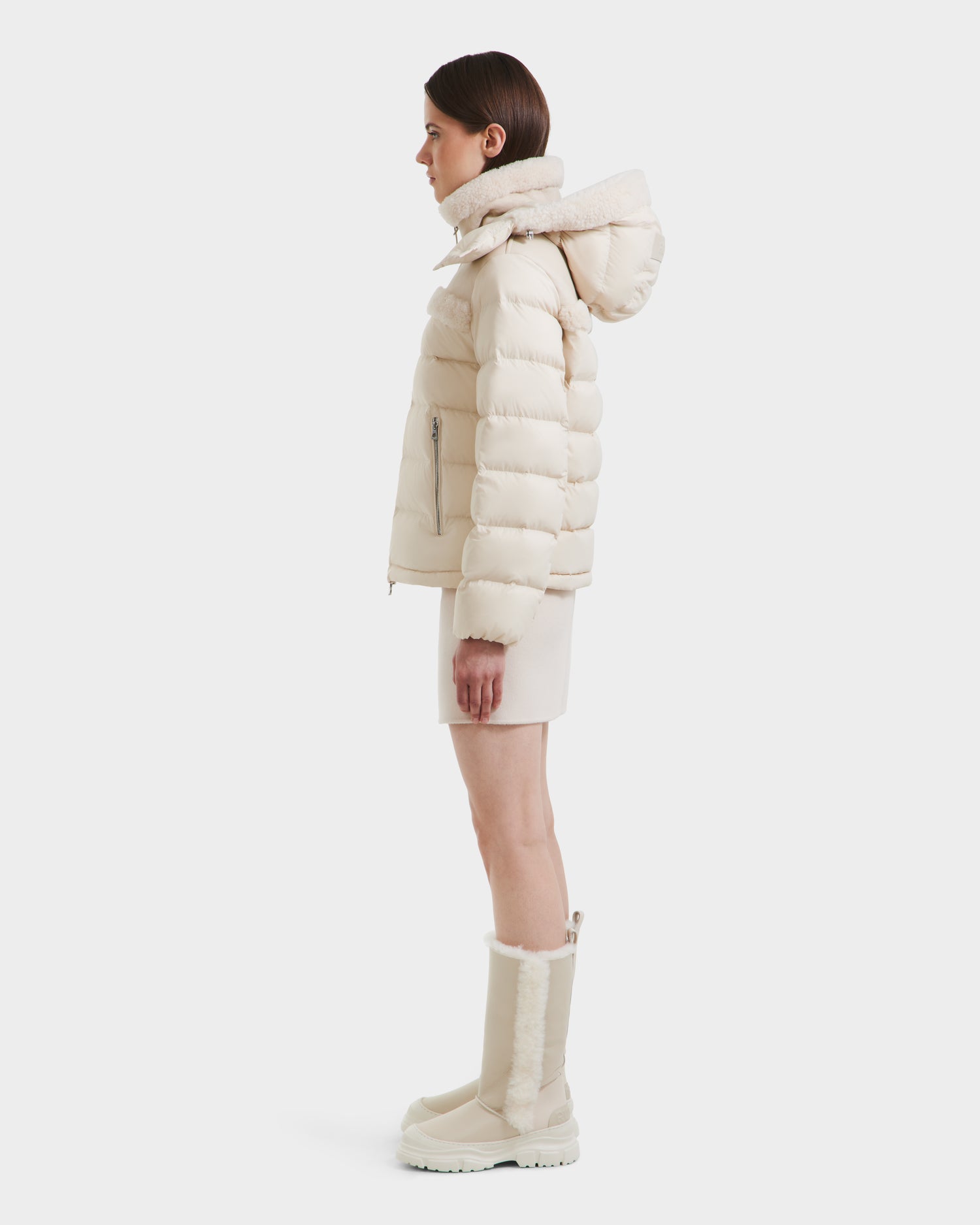 A woman stands in profile wearing the Rudsak SANDRA - 8125612 Vanilla women’s shearling puffer jacket with a hood, styled with a short cream skirt and matching knee-high boots against a plain white background.