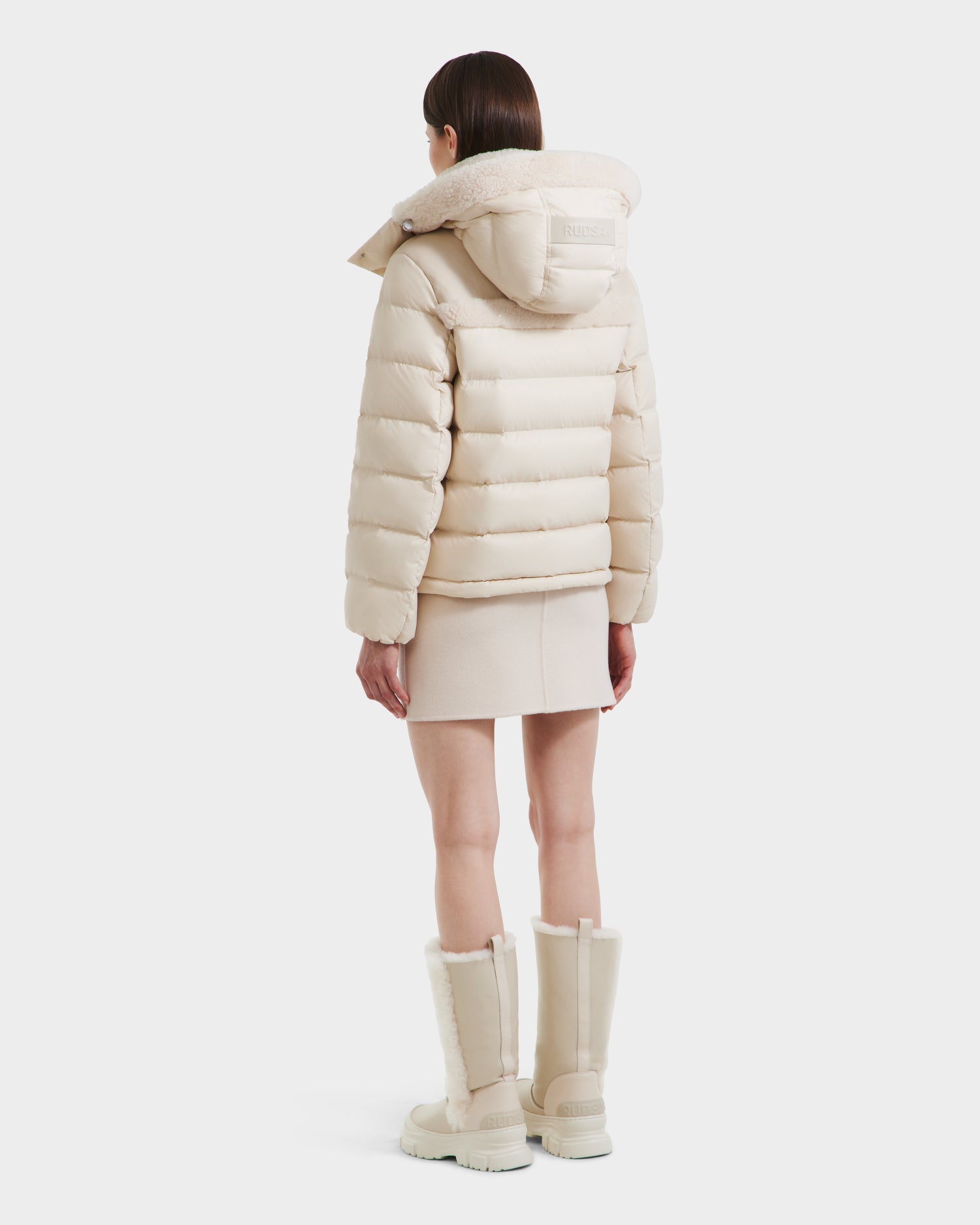 A woman faces away, wearing the Rudsak SANDRA - 8125612 Vanilla women's shearling puffer jacket with hood, styled with a beige mini skirt and fur-trimmed knee-high boots, set against a plain white background.