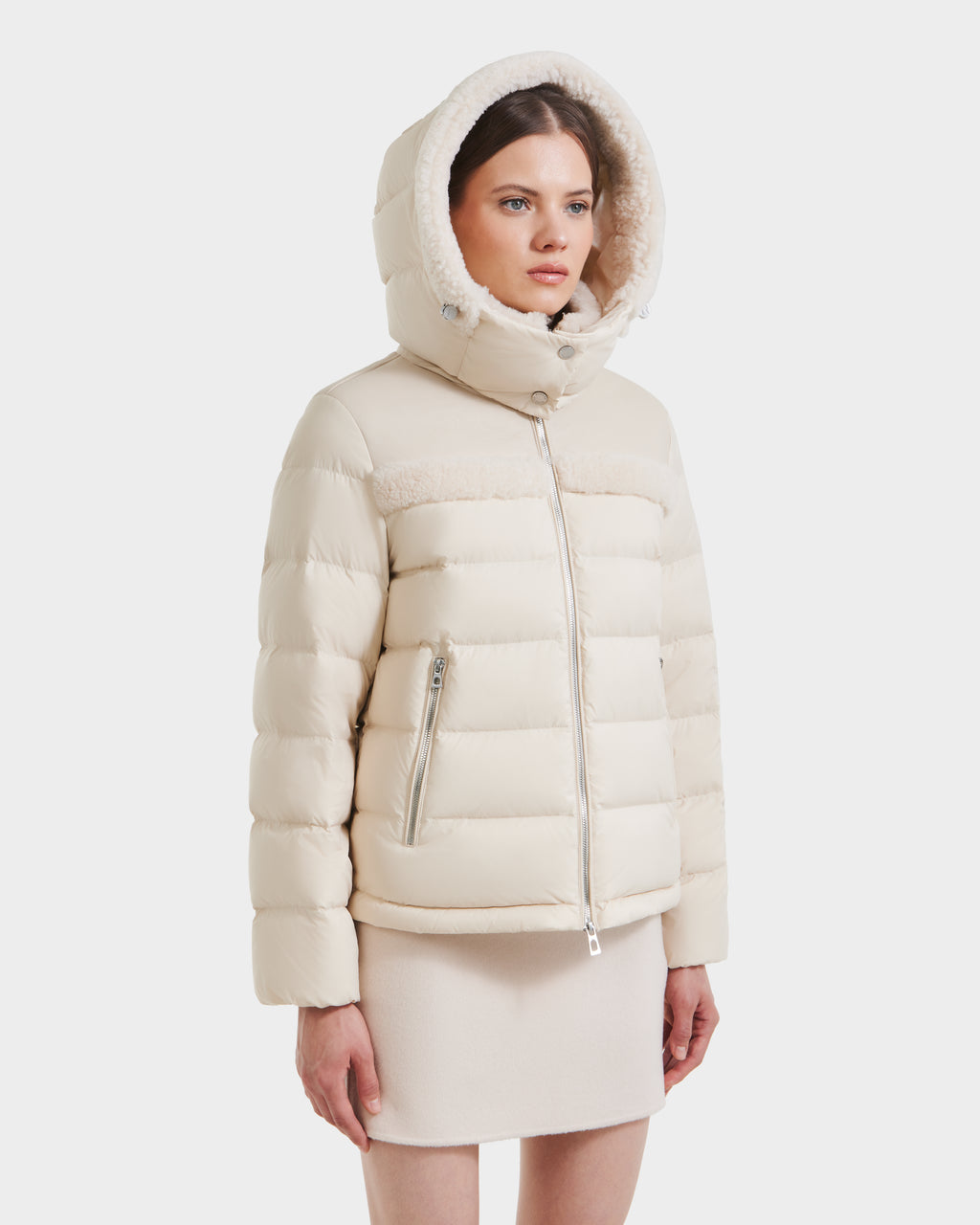A woman wears the Rudsak SANDRA - 8125612 Vanilla puffer jacket with a recycled nylon shell and faux fur-lined hood, paired with a beige skirt, standing against a plain white background.