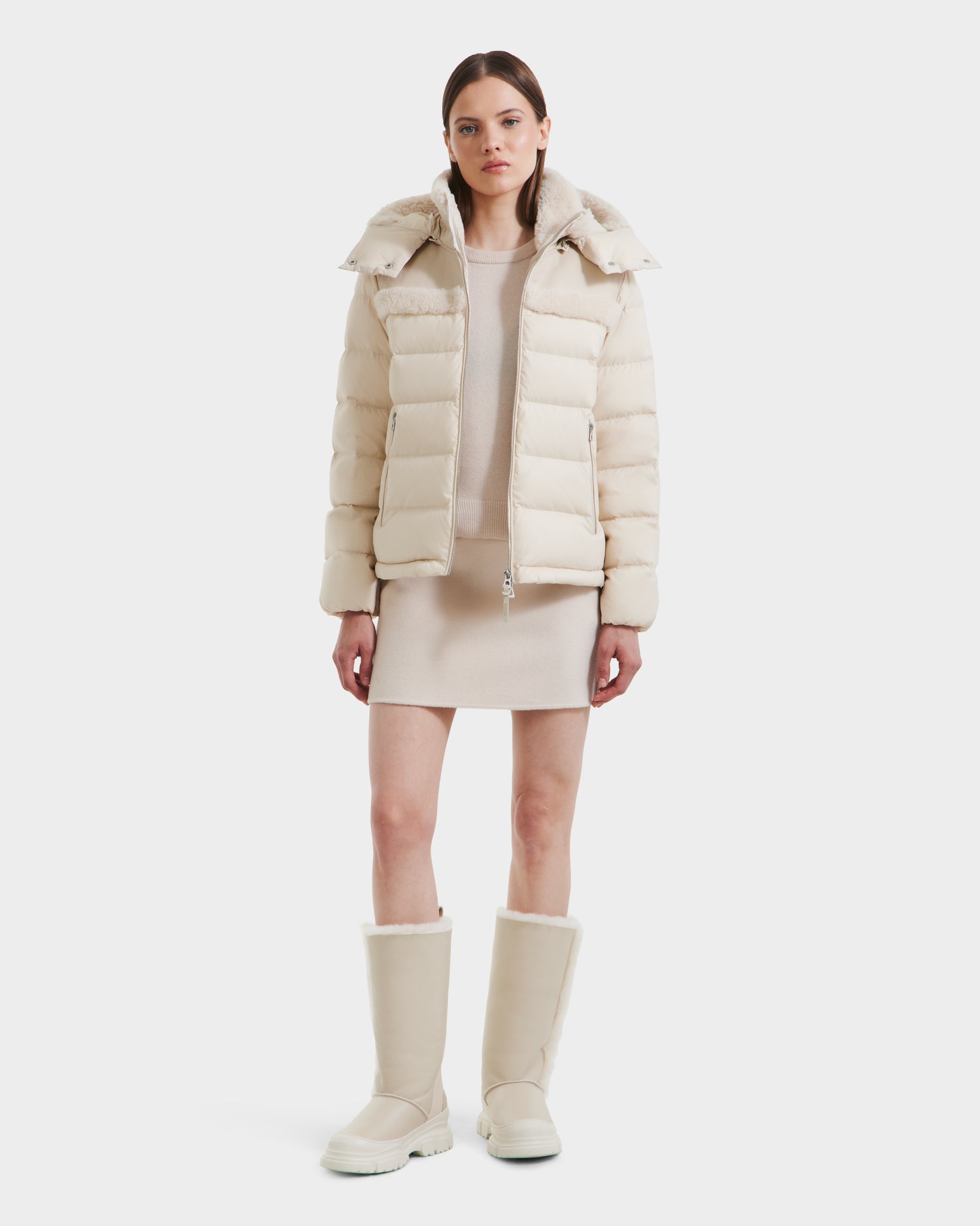A woman with straight brown hair and a neutral expression wears the Rudsak SANDRA - 8125612 Vanilla recycled nylon jacket over a matching dress and tall cream boots, standing against a plain white background.