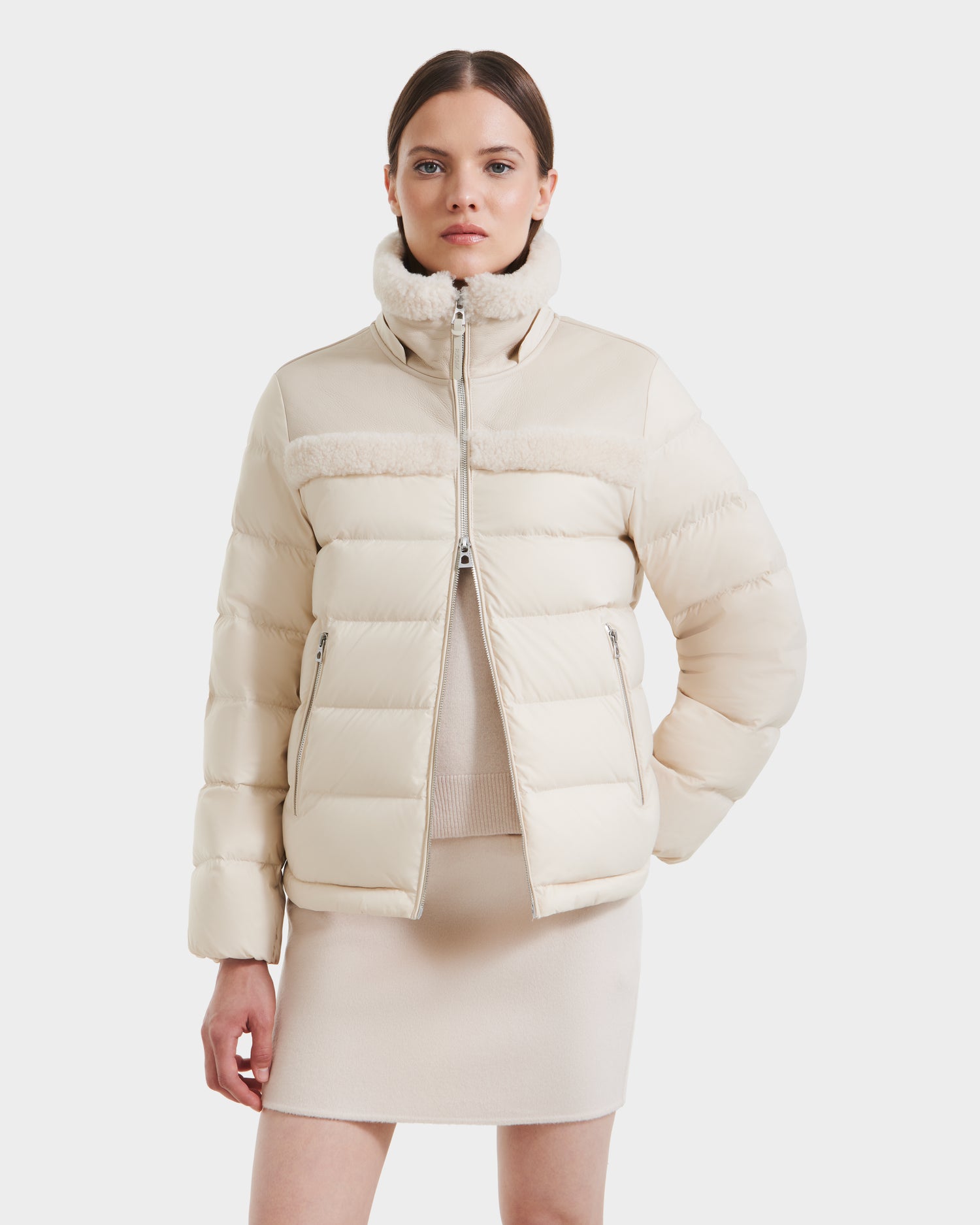 A woman wearing the Rudsak SANDRA - 8125612 Vanilla shearling puffer jacket and a matching cream mini skirt stands against a white background, looking at the camera with her hands in her pockets.