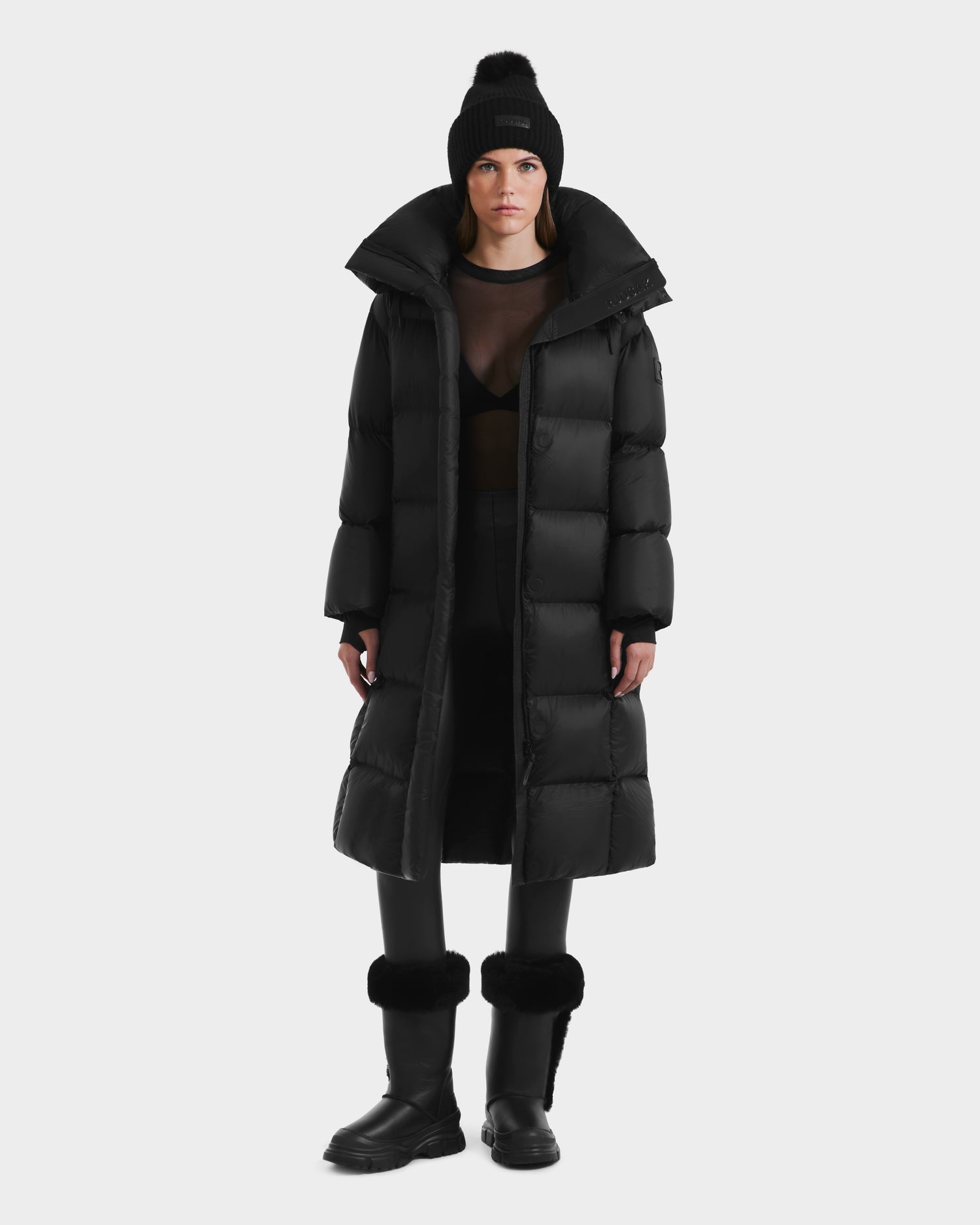 A person wears the Rudsak SASHA - 8125686 Black womens long down puffer coat with removable hood, styled with a black beanie, sheer black top, black pants, and boots, standing facing forward against a plain white background.
