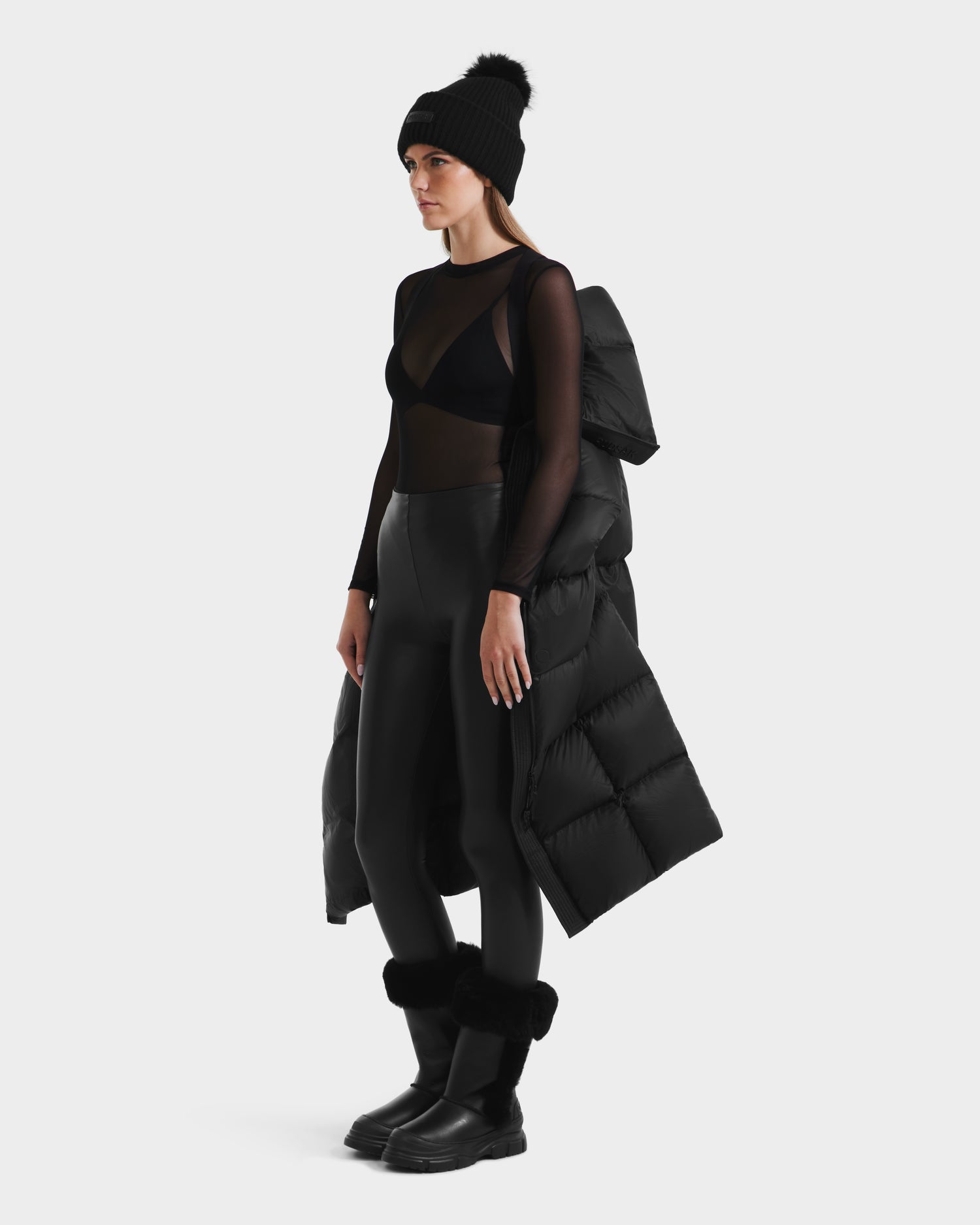 A woman poses against a white background wearing the Rudsak SASHA - 8125686 Black womens long down puffer coat with removable hood draped over her shoulders, paired with black boots, a beanie, sheer top, bra, and high-waisted leggings.