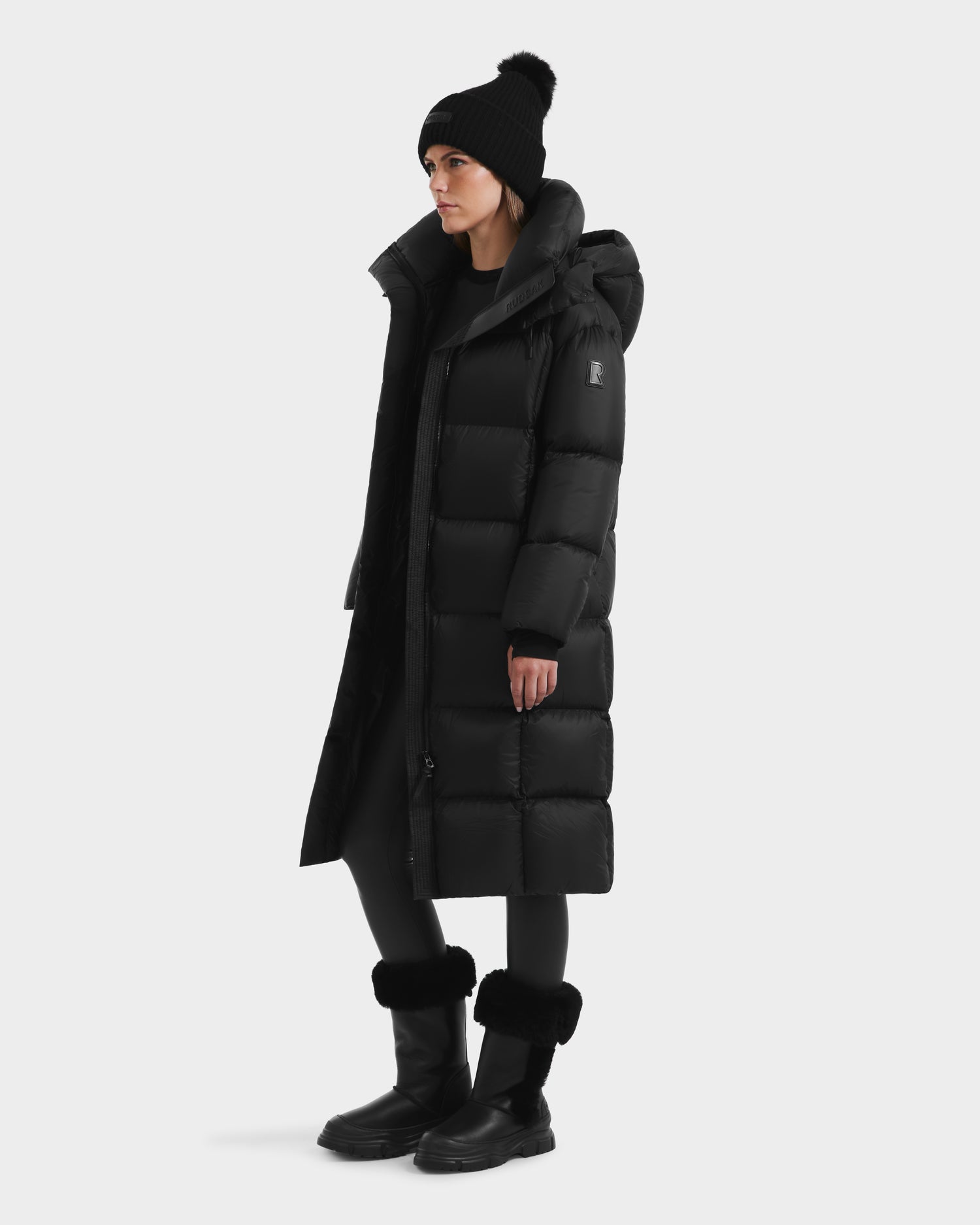 A person in the Rudsak SASHA - 8125686 Black womens long down puffer coat with removable hood, black leggings, fur-trimmed tall black boots, and a pom-pom beanie stands against a plain white background.