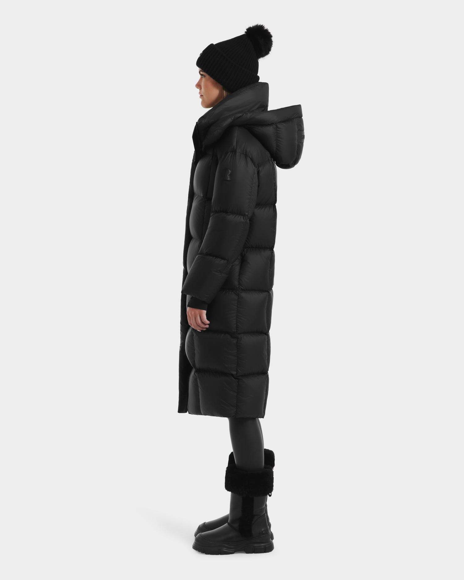 A person stands in profile wearing the Rudsak SASHA - 8125686 Black long down puffer coat with removable hood, paired with a black pom-pom beanie, scarf, leggings, and fur-cuffed boots against a plain light background.