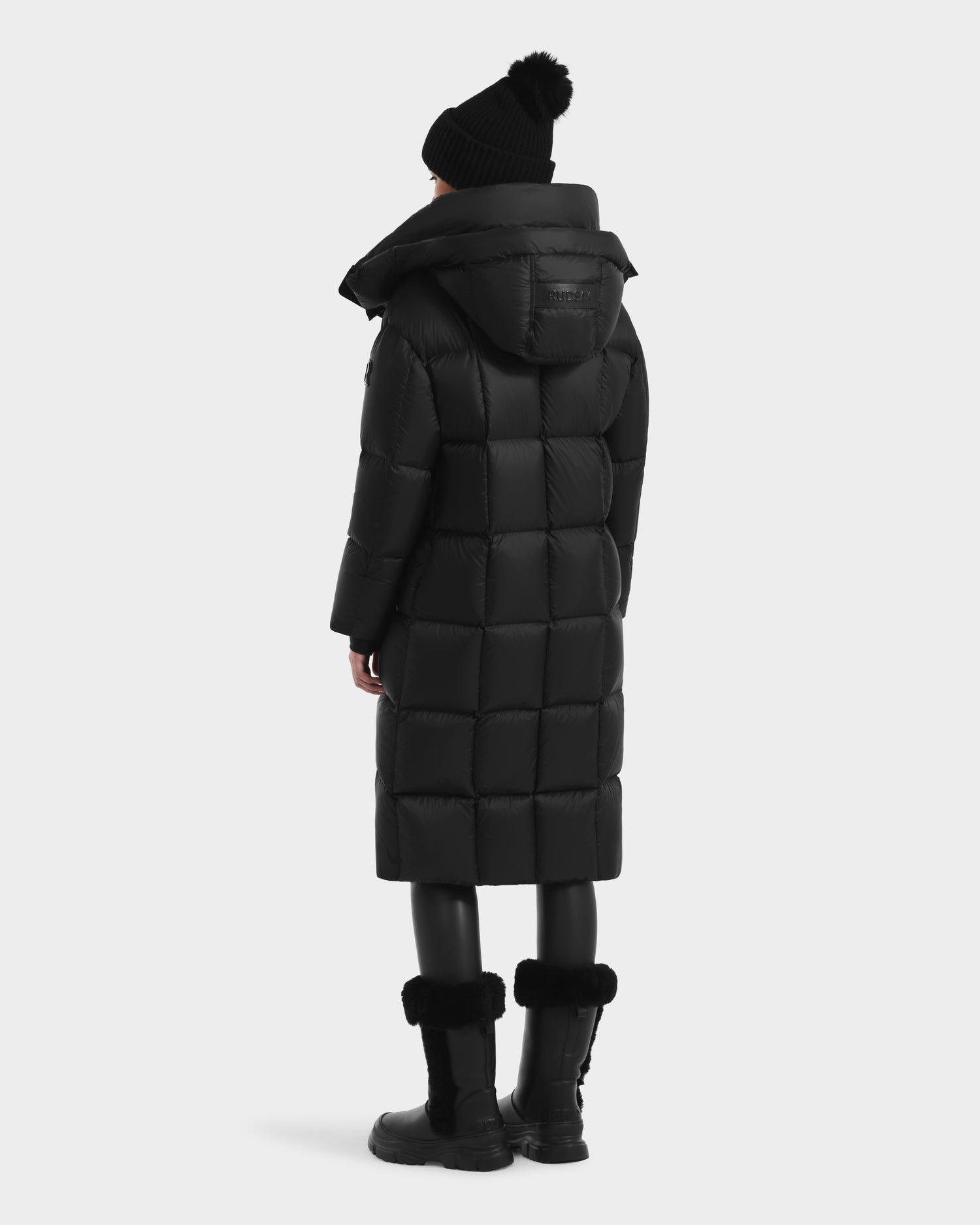 A person wearing the Rudsak SASHA - 8125686 Black women’s long down puffer coat with removable hood, a black pom-pom beanie, black leggings, and fur-trimmed boots stands facing away from the camera against a plain background.