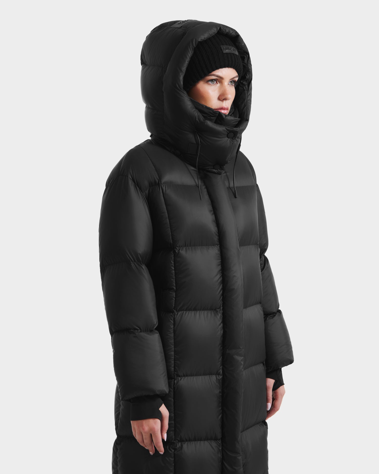 A person wearing the Rudsak SASHA - 8125686 Black women’s long down puffer coat with a removable hood and a black knit beanie stands facing slightly to the right against a plain white background.