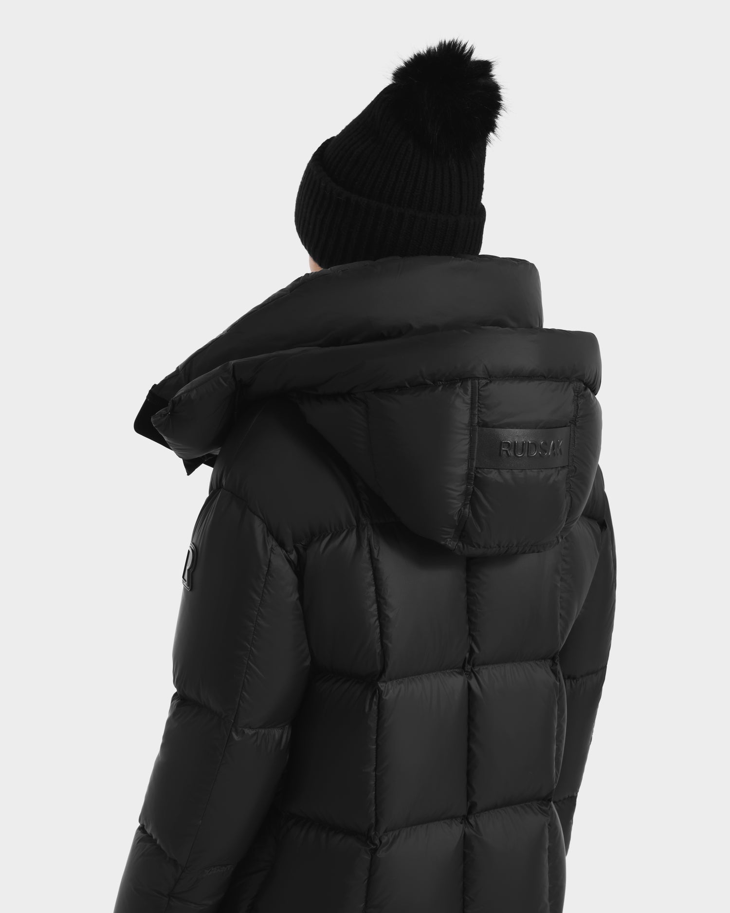 A person is shown from the back in a Rudsak SASHA - 8125686 Black womens long down puffer coat with a large removable hood, paired with a black knit pom-pom beanie, against a plain light background.