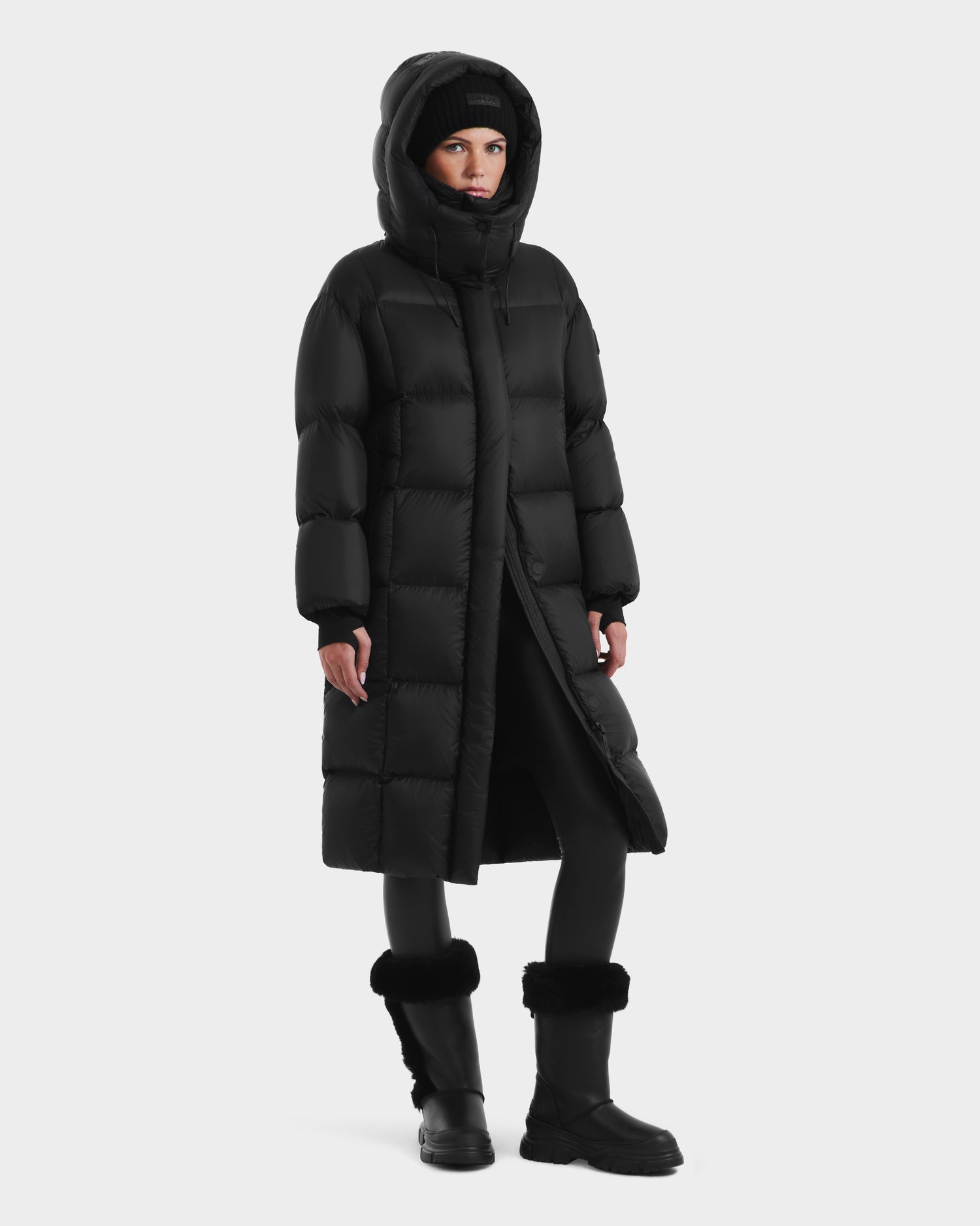 A person wearing the Rudsak SASHA - 8125686 Black women’s long down puffer coat with a removable hood, paired with black leggings and fur-trimmed black winter boots, stands against a plain white background.