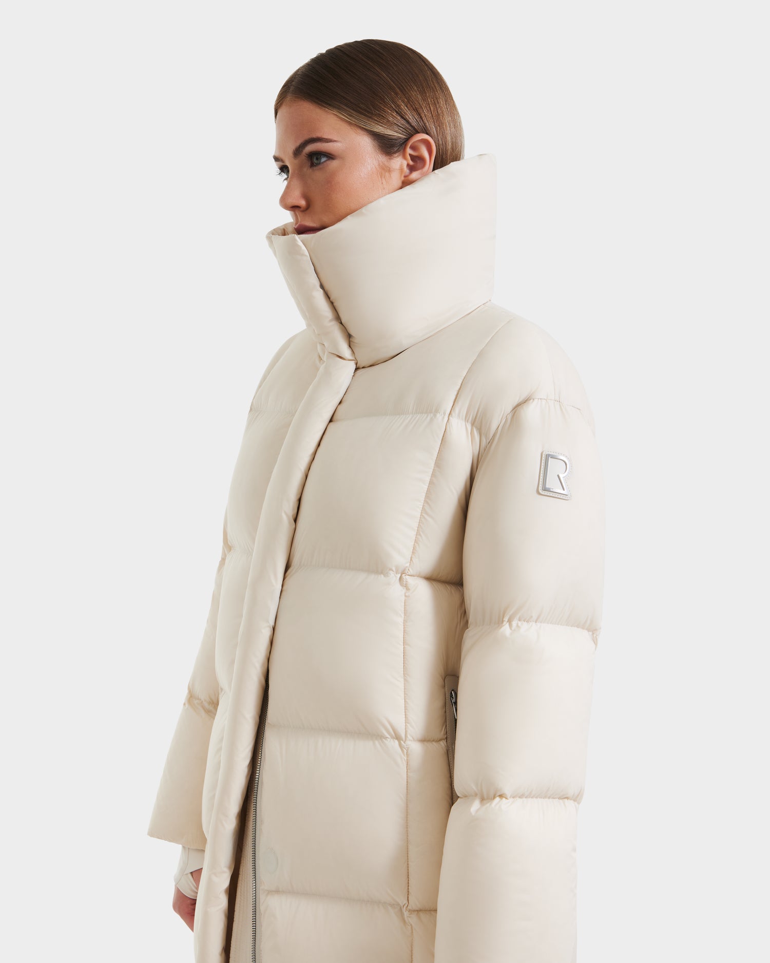 A woman wears the Rudsak SASHA - 8125686 Vanilla long down puffer coat with a high collar, quilted stitching, and a sleeve patch. Her hair is tucked behind her ears as she faces left against a plain white background.