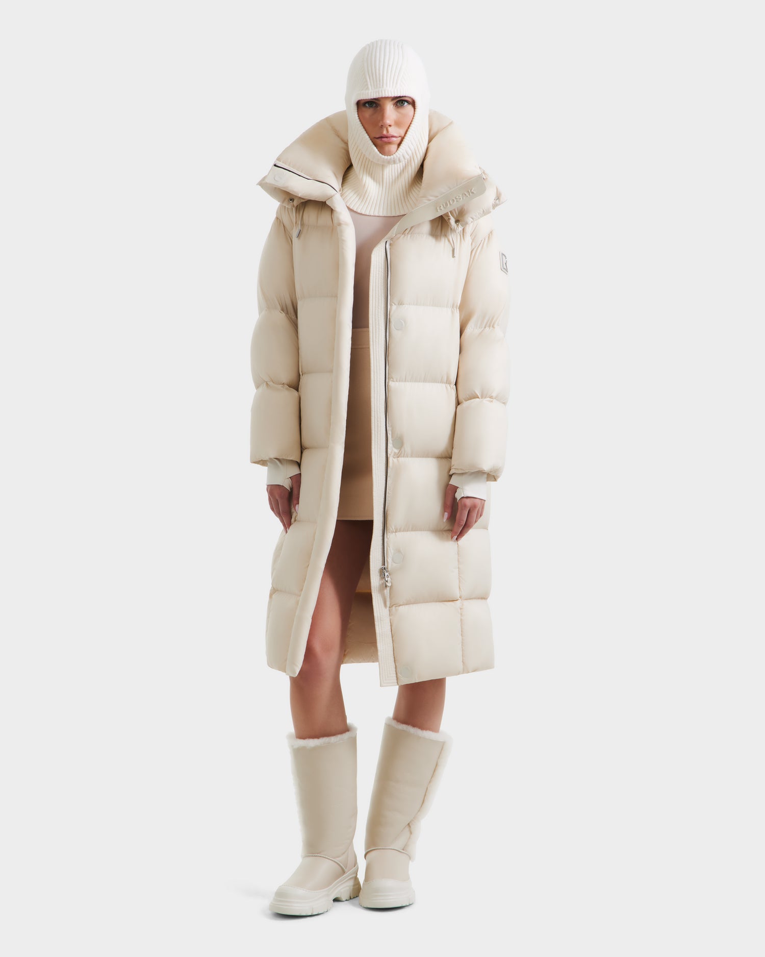 A person faces forward against a plain white background, wearing Rudsaks SASHA - 8125686 Vanilla women’s long down puffer coat with removable hood, paired with a matching knit beanie, turtleneck, and cream mid-calf thick-soled boots.