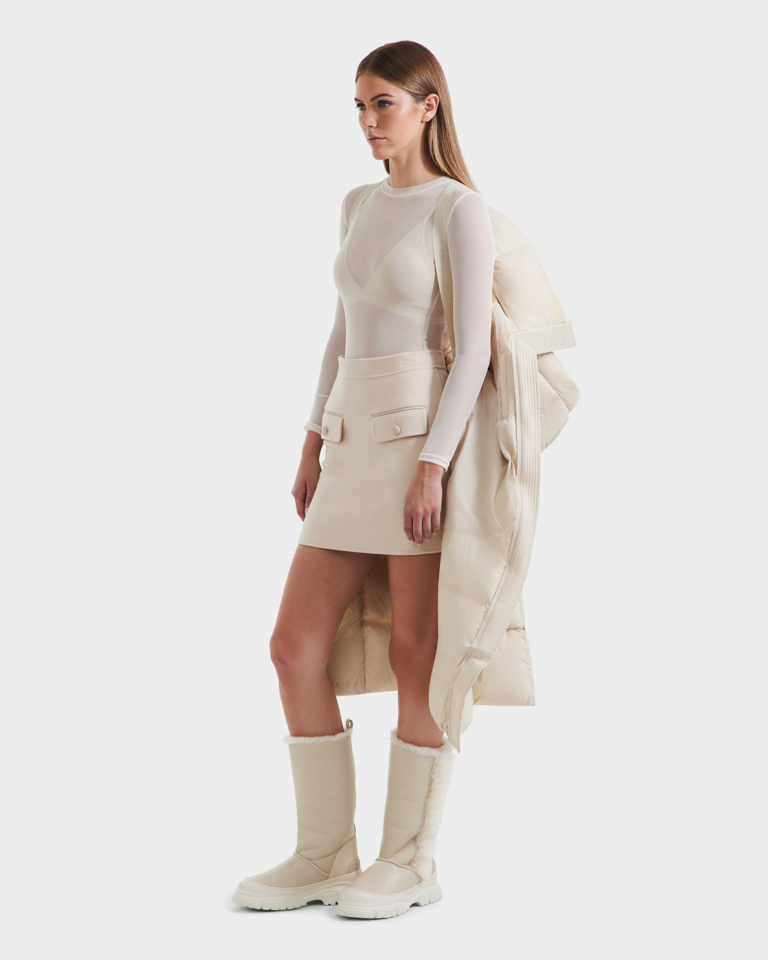 A woman stands against a white background wearing a sheer long-sleeve top, cream mini skirt with pockets, tall white boots, and holding the Rudsak SASHA - 8125686 Vanilla womens long down puffer coat with removable hood over her arm.