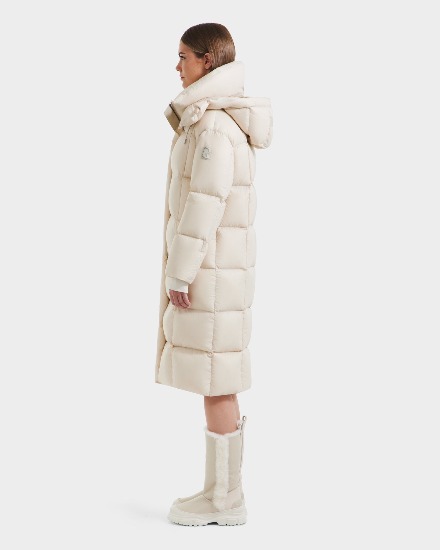 A person stands in profile wearing the Rudsak SASHA - 8125686 Vanilla women’s long down puffer coat with a high collar and removable hood, paired with matching mid-calf winter boots, against a plain white background.