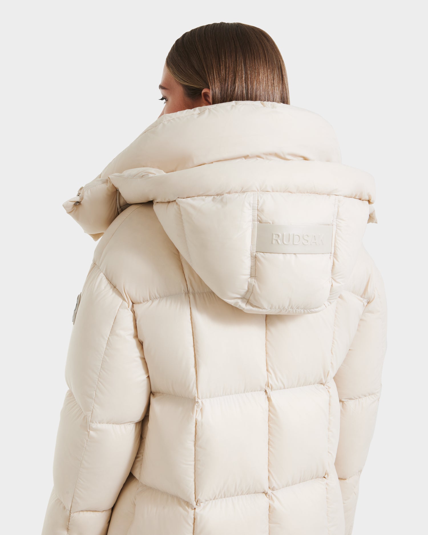 A person with straight brown hair is shown from the back wearing the Rudsak SASHA - 8125686 Vanilla women’s long down puffer coat in pale beige with a large removable hood and brand patch on the back of this waterproof winter coat.