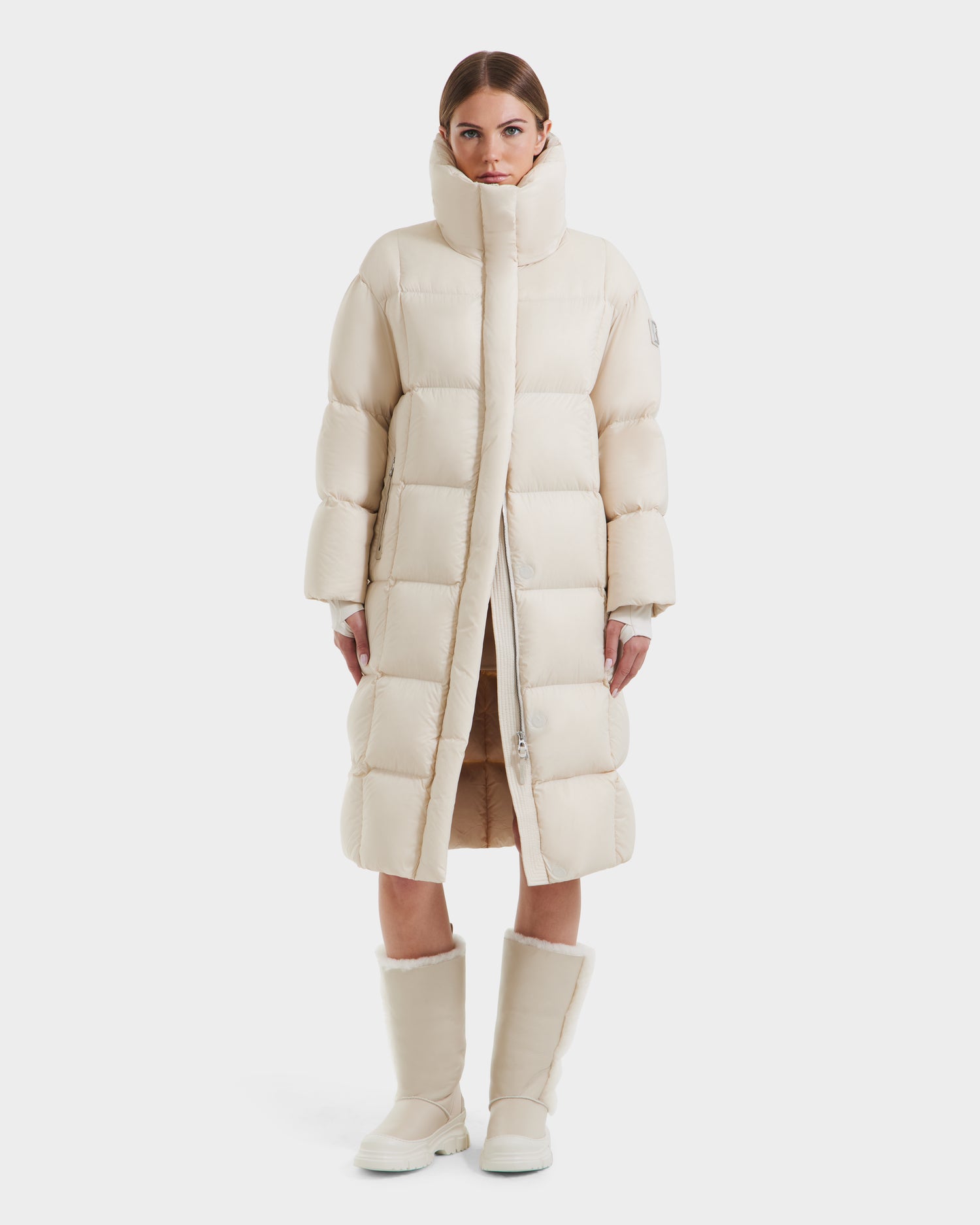 A woman stands facing forward, wearing the Rudsak SASHA - 8125686 Vanilla long down puffer coat with a high collar, removable hood, and matching mid-calf boots. The cream-colored winter coat is set against a plain white background.
