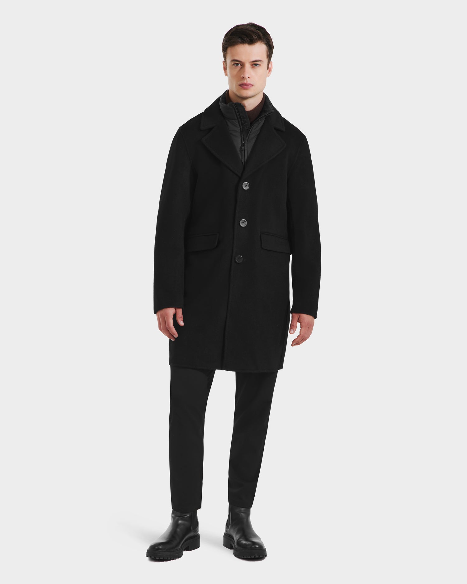A man stands facing forward, wearing the Rudsak SAWYER - 6125511 Black wool blend coat over a black zip-up layer, paired with black pants and boots, posing against a plain white background.