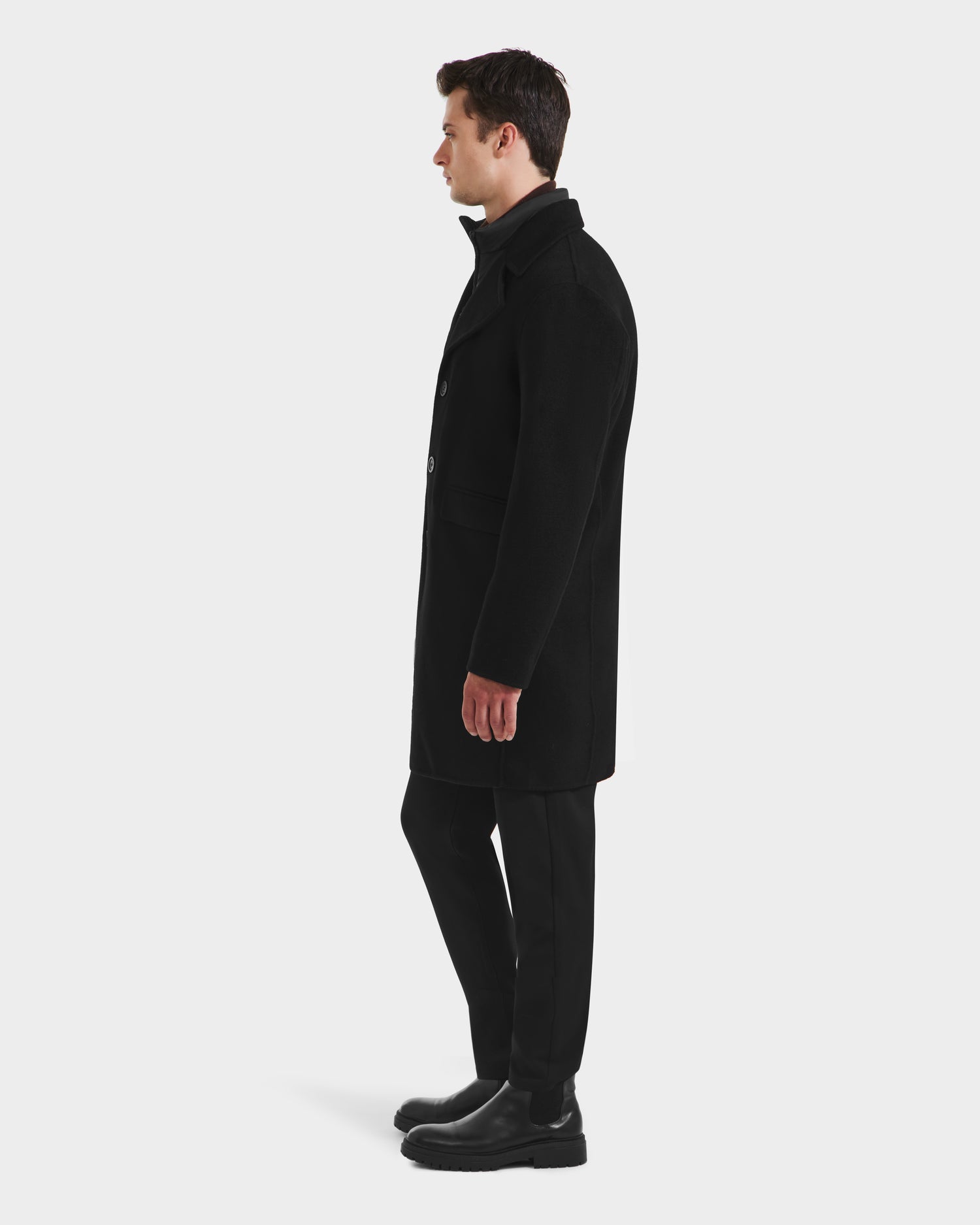 A man stands in profile against a white background, wearing the Rudsak SAWYER - 6125511 Black men’s wool blend coat, black trousers, and black shoes for a classic, elevated outerwear look.