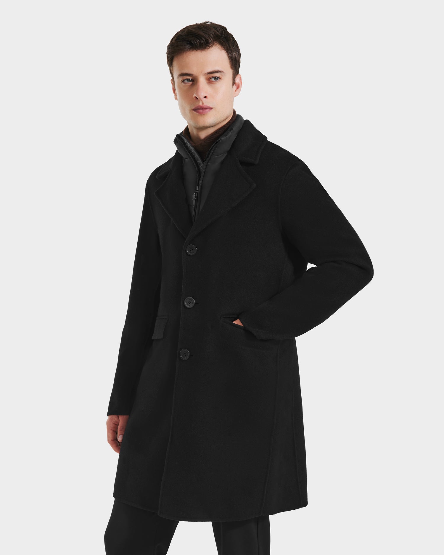 A man wears the Rudsak SAWYER - 6125511 Black mens wool blend coat, featuring three buttons, side pockets, and a removable quilted dickie, as he stands with one hand in his pocket against a plain light background.