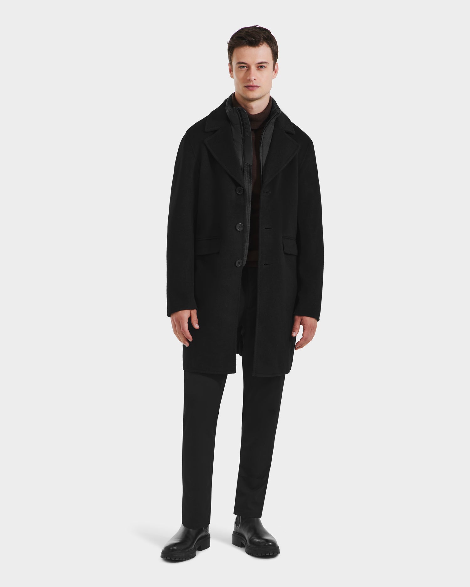 A man wears the Rudsak SAWYER - 6125511 Black wool blend coat over a dark outfit with black pants and shoes, presenting a classic silhouette against a white background.