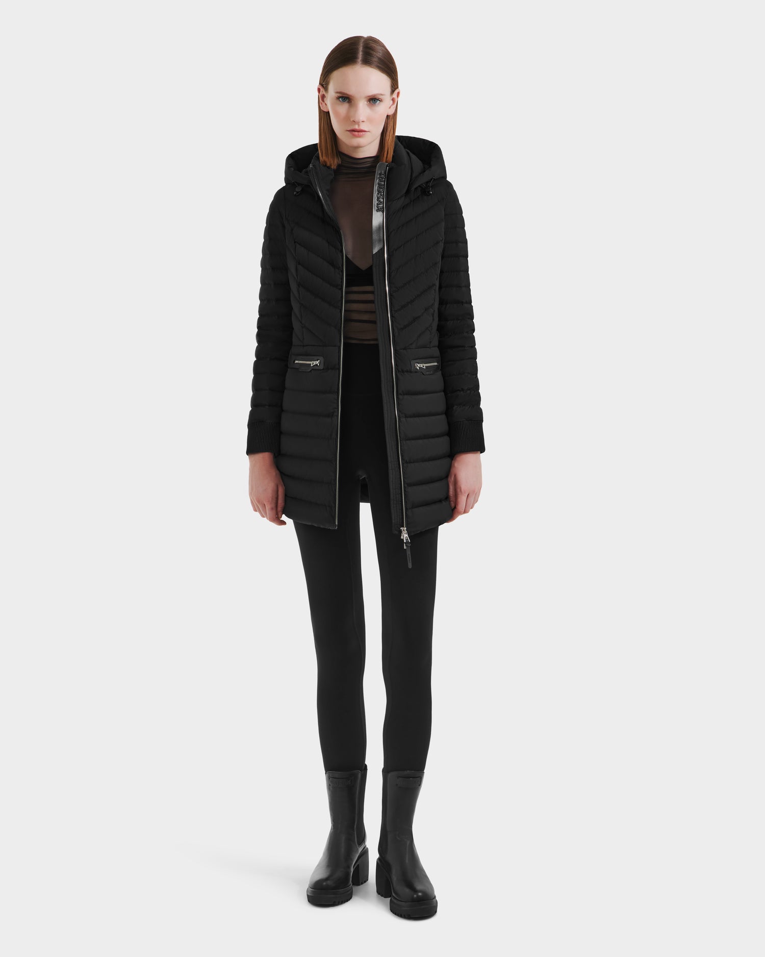 A woman faces forward in a Rudsak SCARLETT M-M - 8125595 Black long quilted coat with hood and zippered pockets, styled with black leggings, boots, and a sheer black top over a bra. The background is plain white.