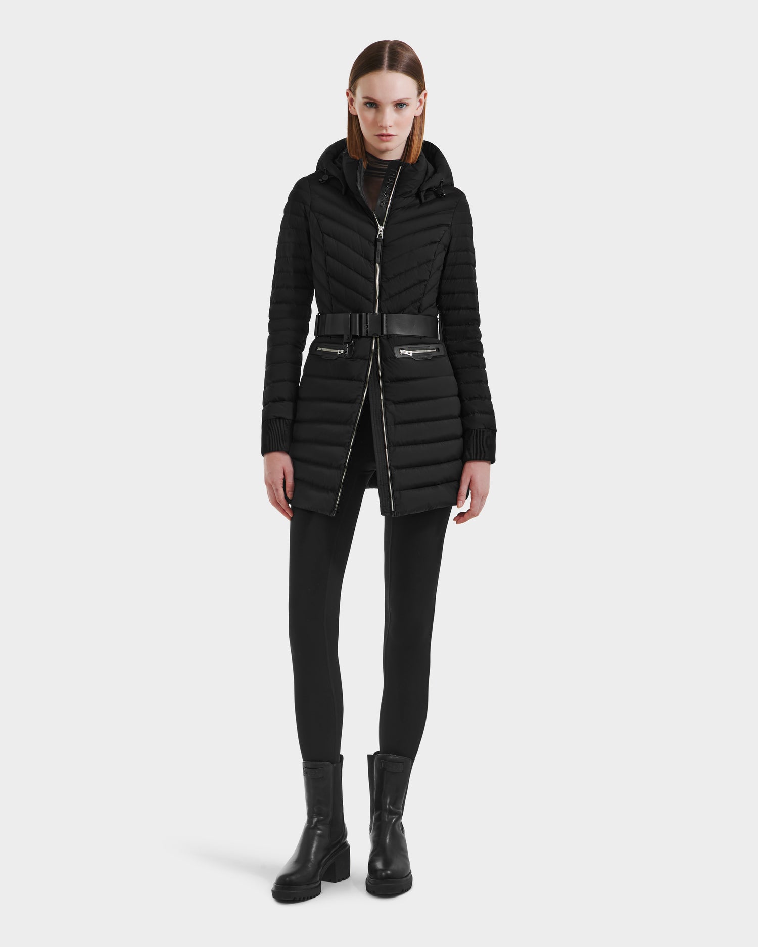 A woman stands facing forward in the Rudsak SCARLETT M-M - 8125595 Black, a mid-length lightweight down puffer coat with a vegan leather belt and zippered pockets, paired with black leggings and ankle boots, against a plain white background.