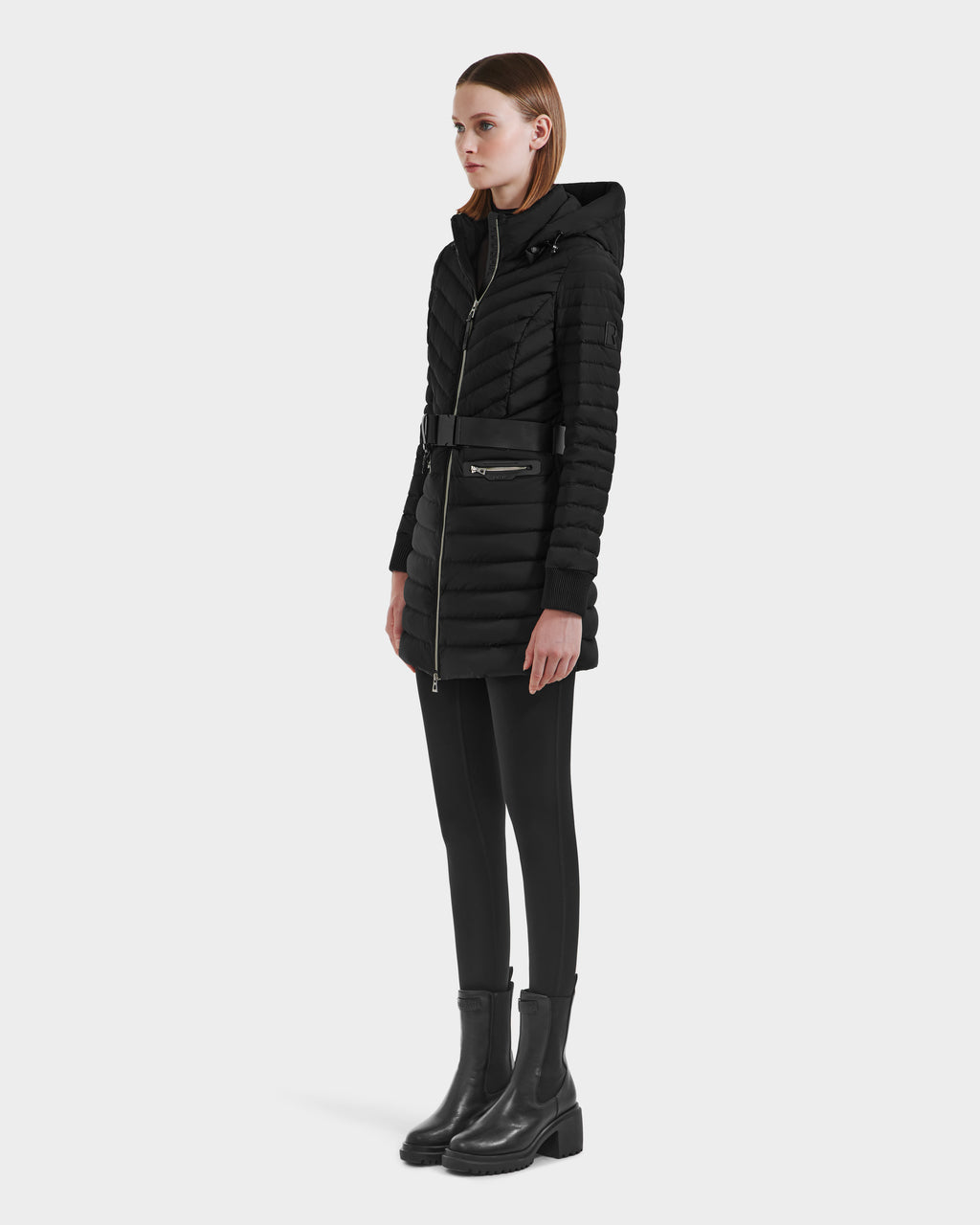 A woman faces slightly to the side in a Rudsak SCARLETT M-M - 8125595 Black lightweight down puffer coat with a vegan leather belt, paired with black leggings and boots, against a plain white background.