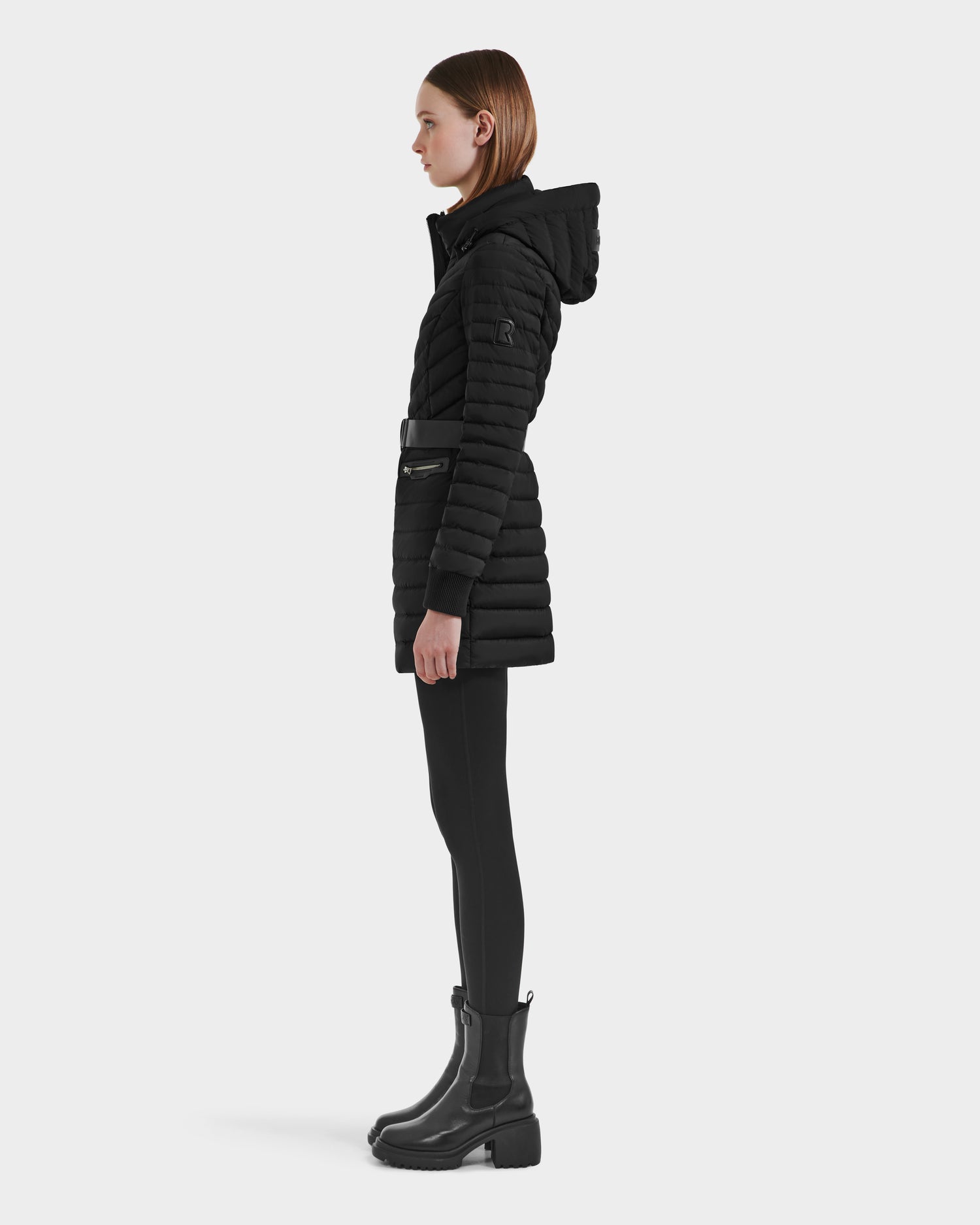A woman stands in profile against a white background wearing the Rudsak SCARLETT M-M - 8125595 Black quilted mid-length hooded coat with a vegan leather belt, paired with black leggings and black ankle boots.