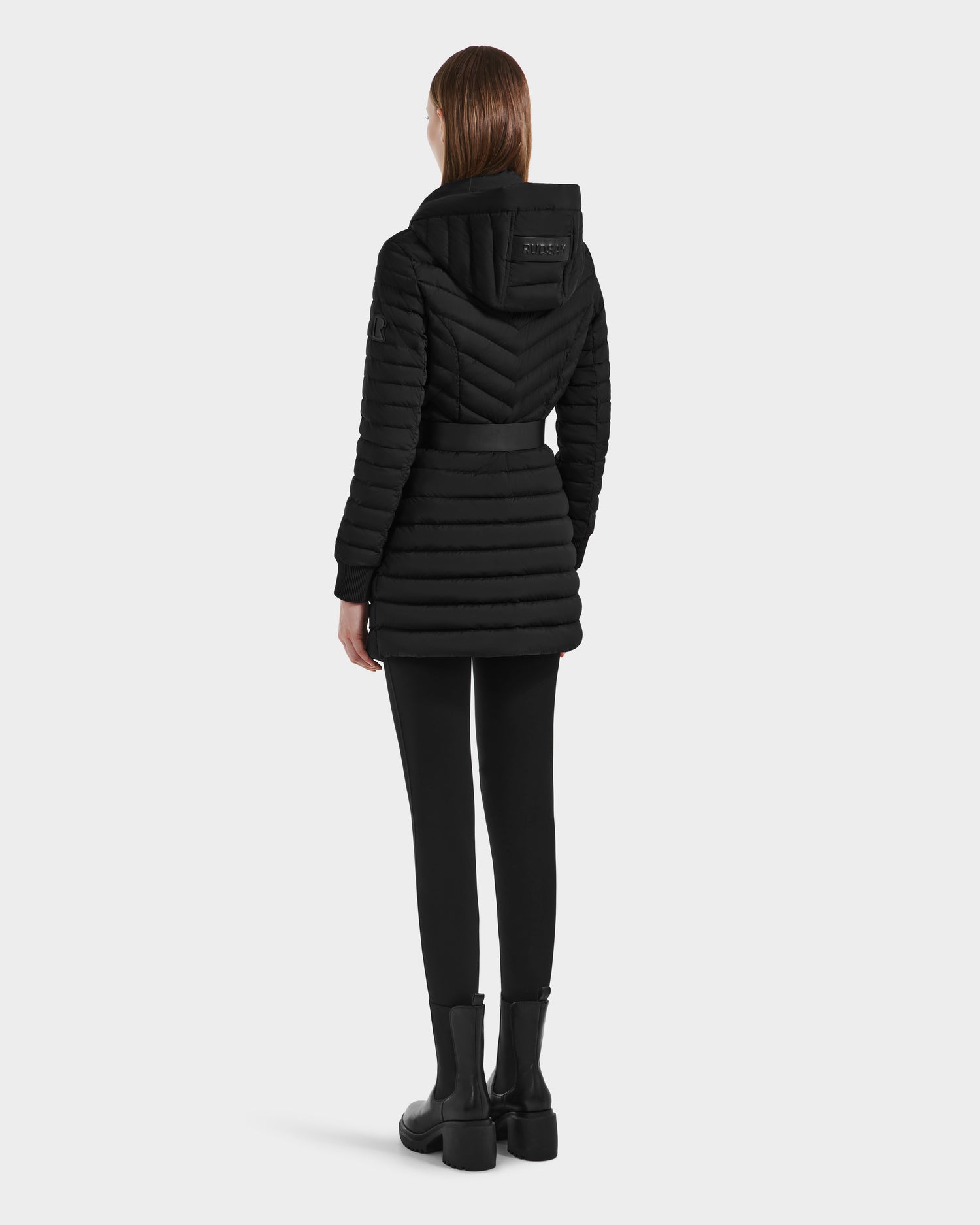 A woman with straight brown hair faces away, wearing the Rudsak SCARLETT M-M - 8125595 Black lightweight down puffer with quilted hood, paired with black leggings and ankle boots against a light gray background.