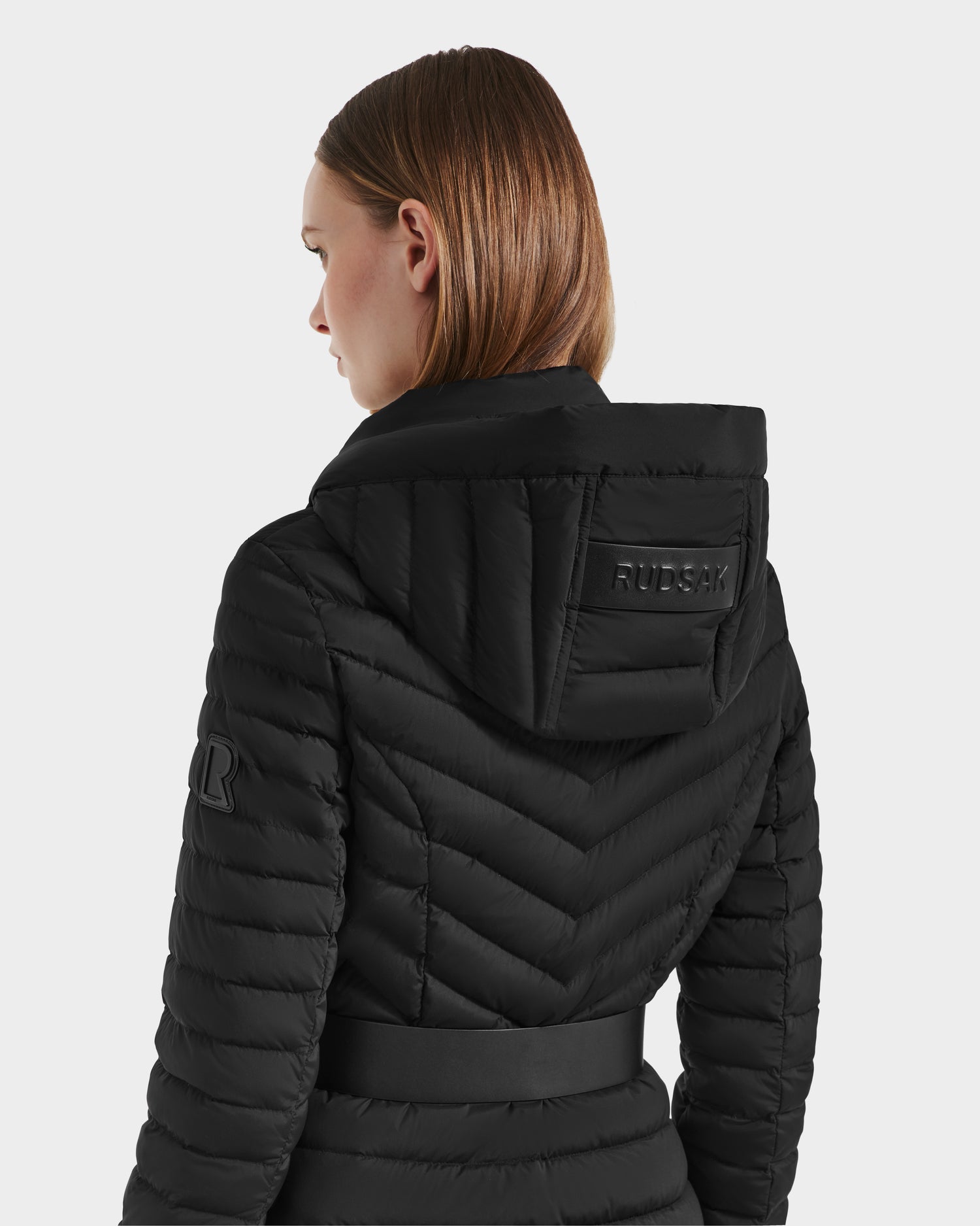 A woman with straight light brown hair wears the Rudsak SCARLETT M-M - 8125595 Black puffer jacket, featuring a hood, vegan leather belt, RUDSAK patch on the hood, and logo patch on the sleeve. She faces away from the camera.