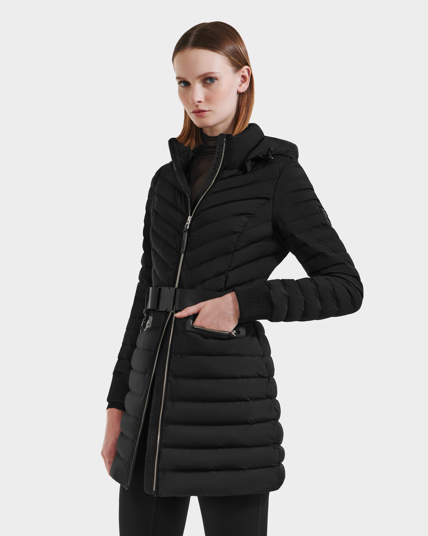 A woman models the Rudsak SCARLETT M-M - 8125595 Black mid-length quilted puffer coat with a hood and vegan leather belt against a light gray background, her straight shoulder-length hair framing a neutral expression.