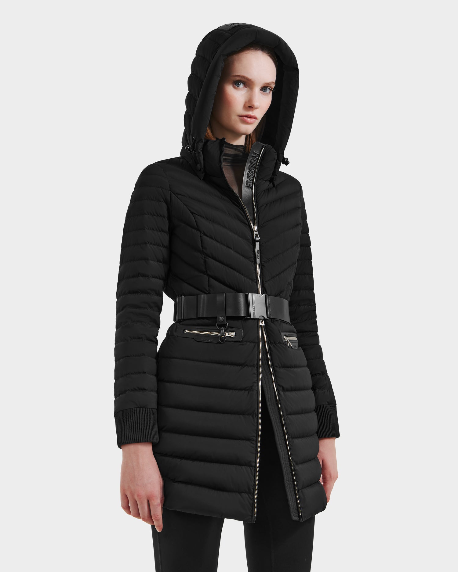 A woman stands against a light gray background wearing the Rudsak SCARLETT M-M - 8125595 Black puffer coat, featuring a zippered front, hood, zippered pockets, and a vegan leather belt.