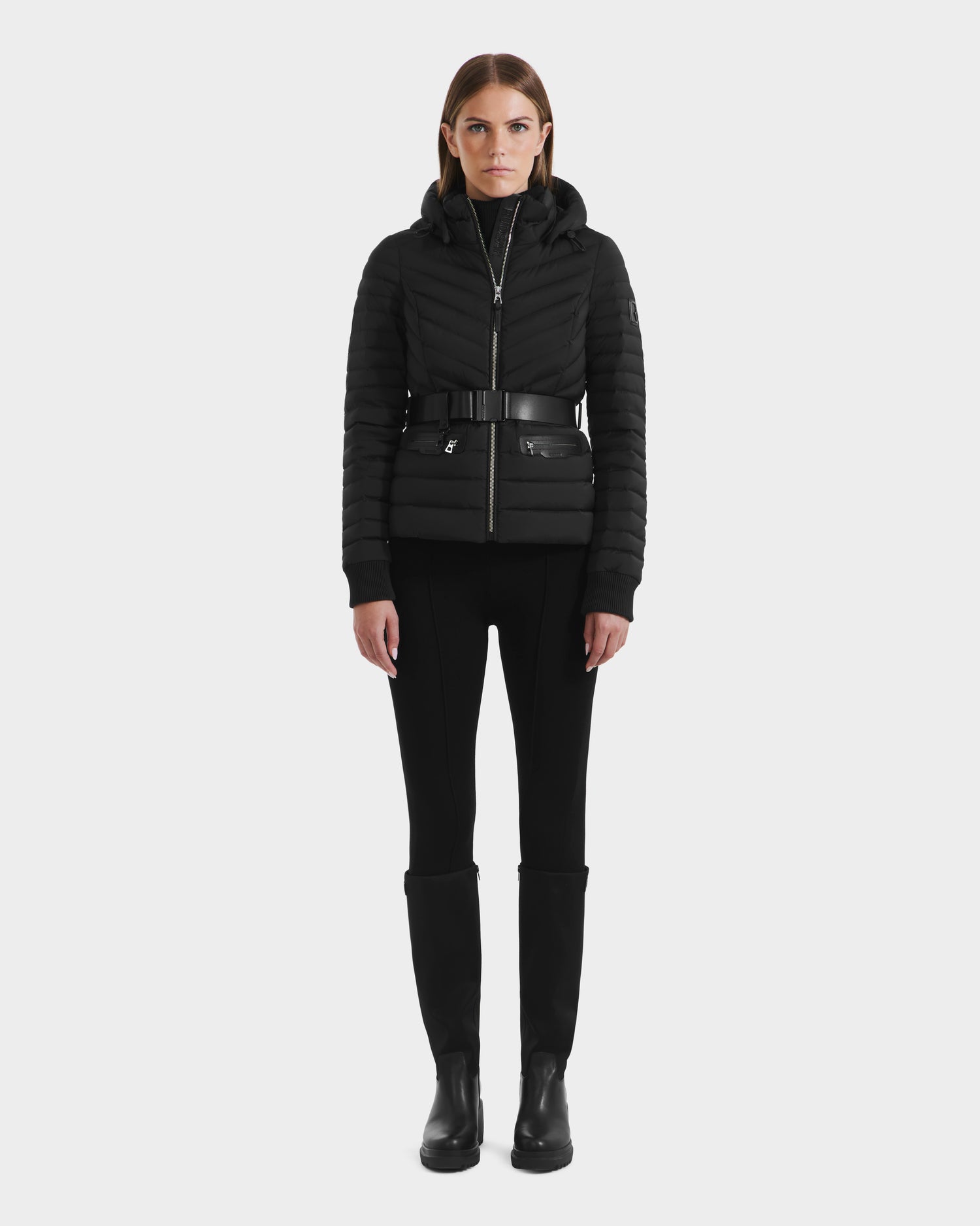 A woman faces forward wearing the Rudsak SCARLETT M - 8125565 Black womens lightweight down jacket with a belted waist, paired with black pants and boots, set against a plain light background.