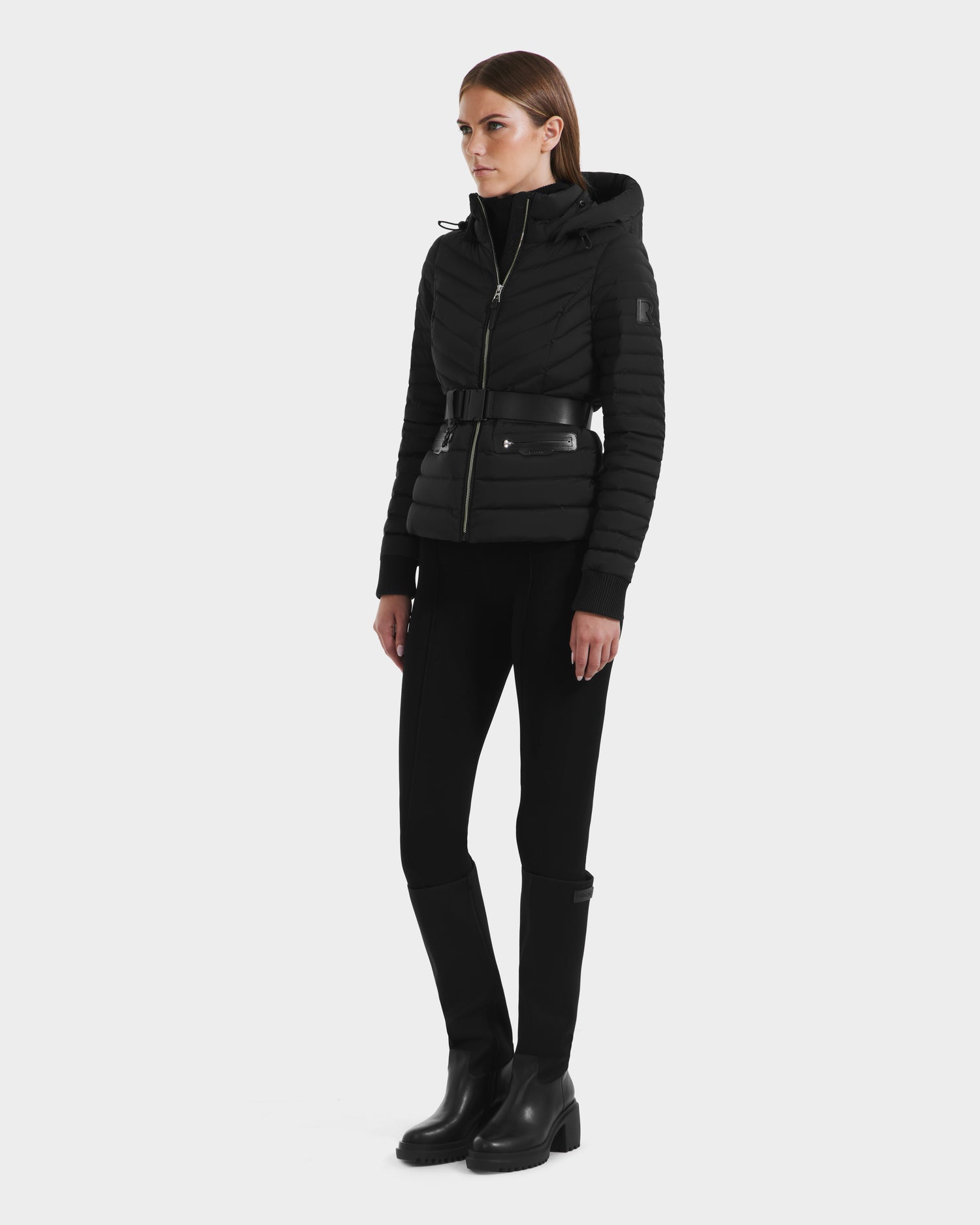 A woman with straight brown hair and a neutral expression stands slightly to the side against a white background, wearing the Rudsak SCARLETT M - 8125565 Black vegan leather belted jacket, paired with black pants and knee-high boots.