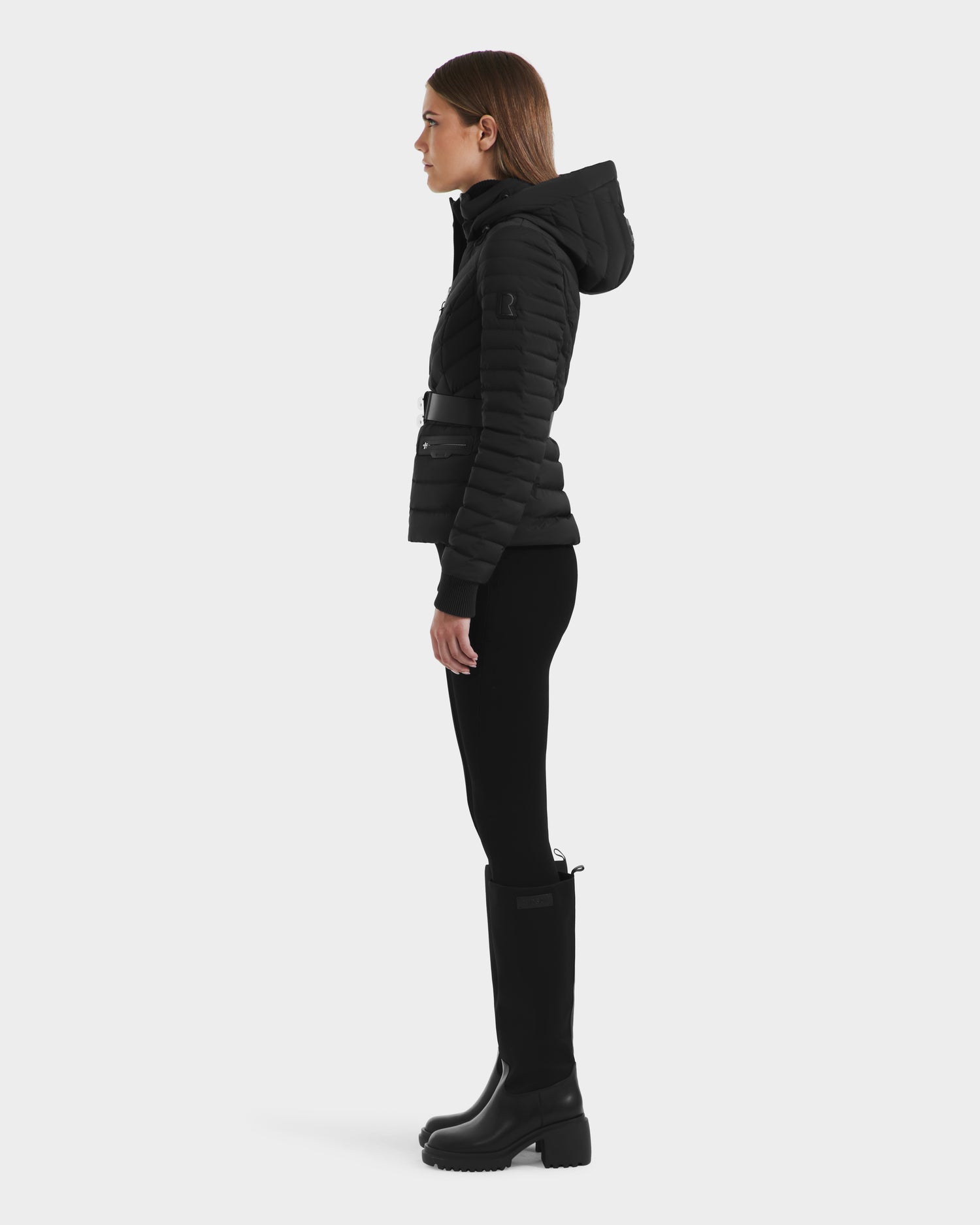 A woman stands in profile against a plain background, wearing the Rudsak SCARLETT M - 8125565 Black lightweight down jacket, paired with black leggings and tall black boots.