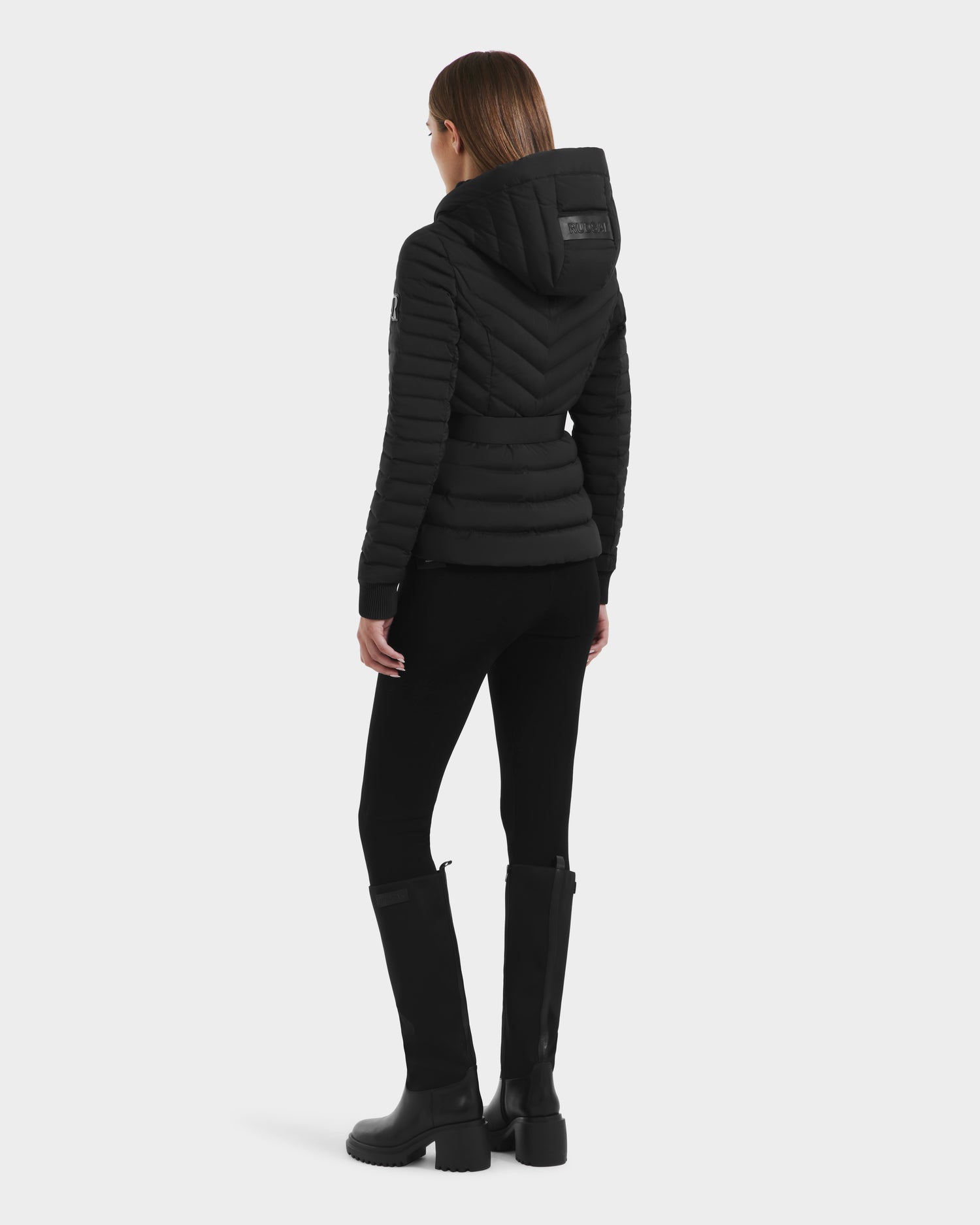A woman faces away, wearing the Rudsak SCARLETT M - 8125565 Black lightweight down jacket with hood, paired with black pants and knee-high boots against a plain light background.