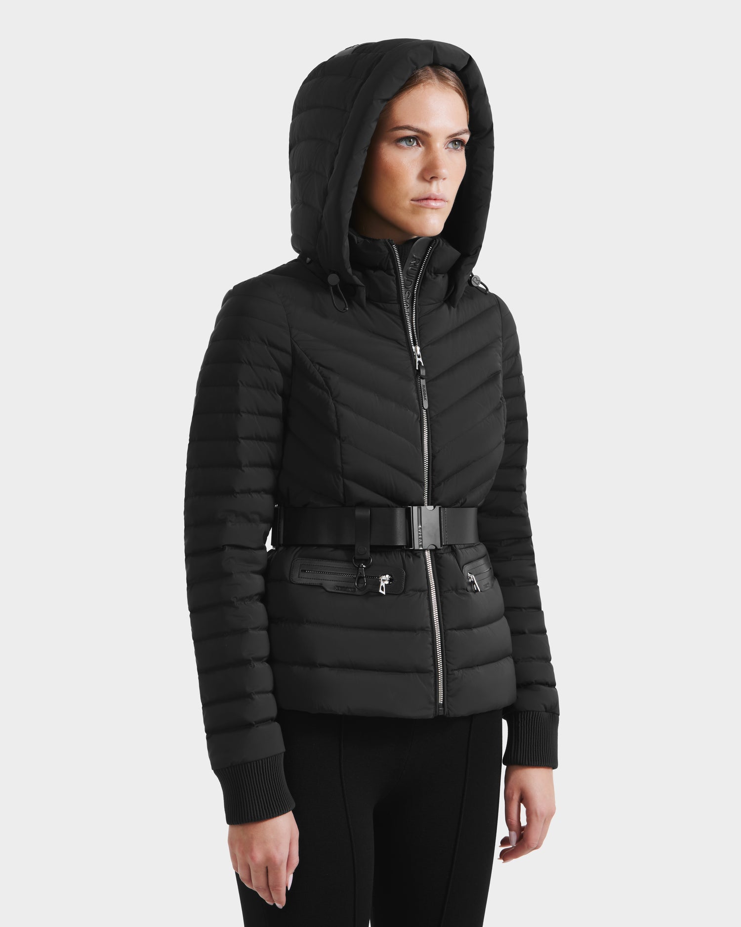 A woman wearing the Rudsak SCARLETT M - 8125565 Black quilted hooded women’s lightweight down jacket with a front zipper, vegan leather belt, and zippered pockets stands against a plain light background.